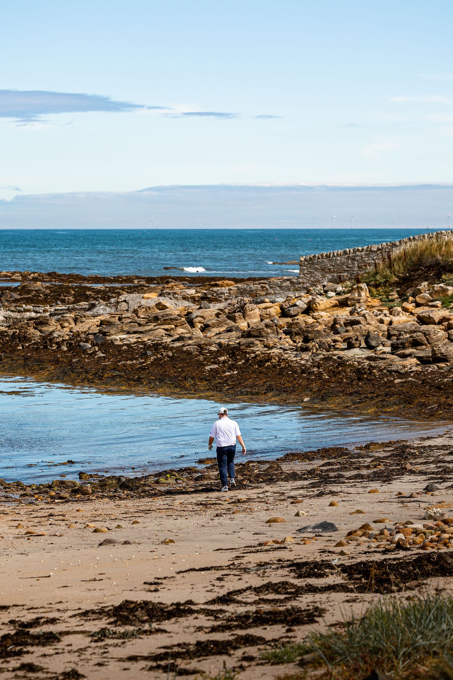 Lone golfer walks along a rocky coastal beach with blue ocean and clear sky stretching into the distance.