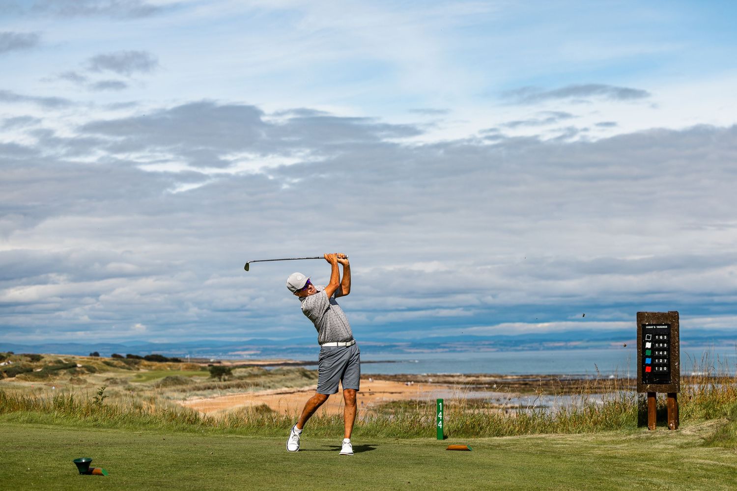 A golfer completes a powerful tee shot on a seaside links course under a dramatic cloudy sky.