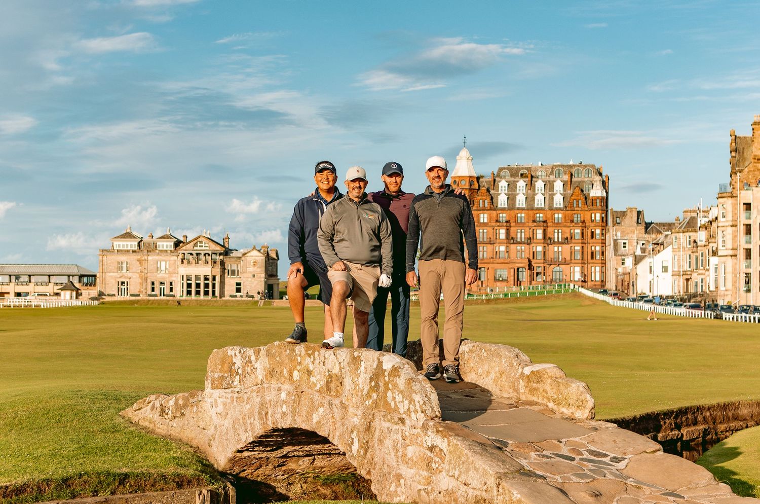 Golfers standing on Swilcan Bridge at St Andrews golf course, Scotland, under blue skies.