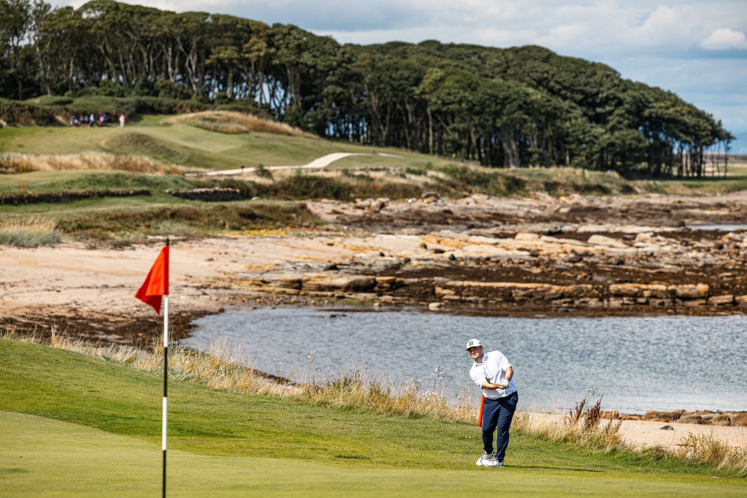 Golfer chips toward the pin on a coastal links course with a red flag, beach, and rocky shoreline nearby.