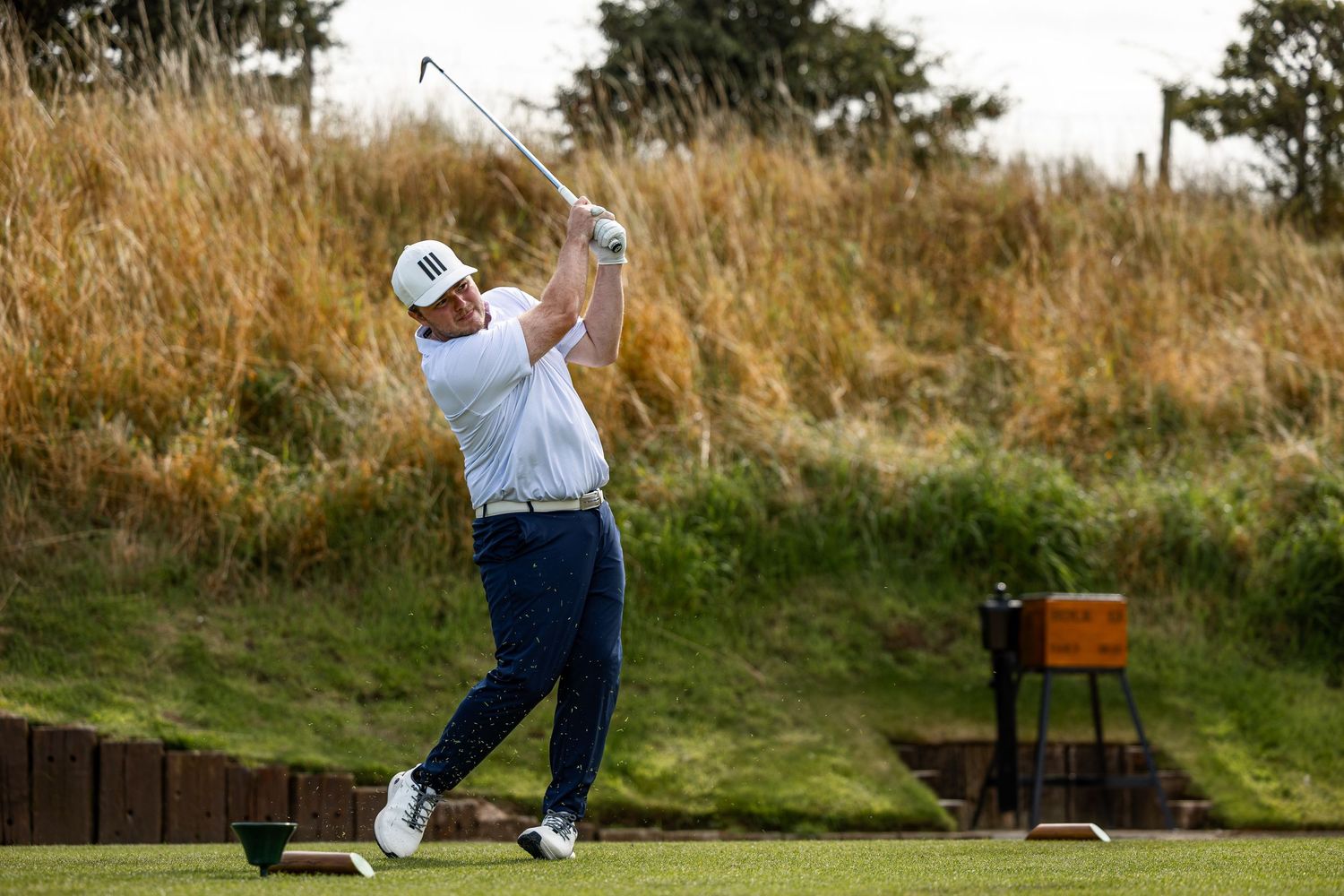 Golfer completes a full swing on a links course surrounded by tall golden rough grass under cloudy skies.