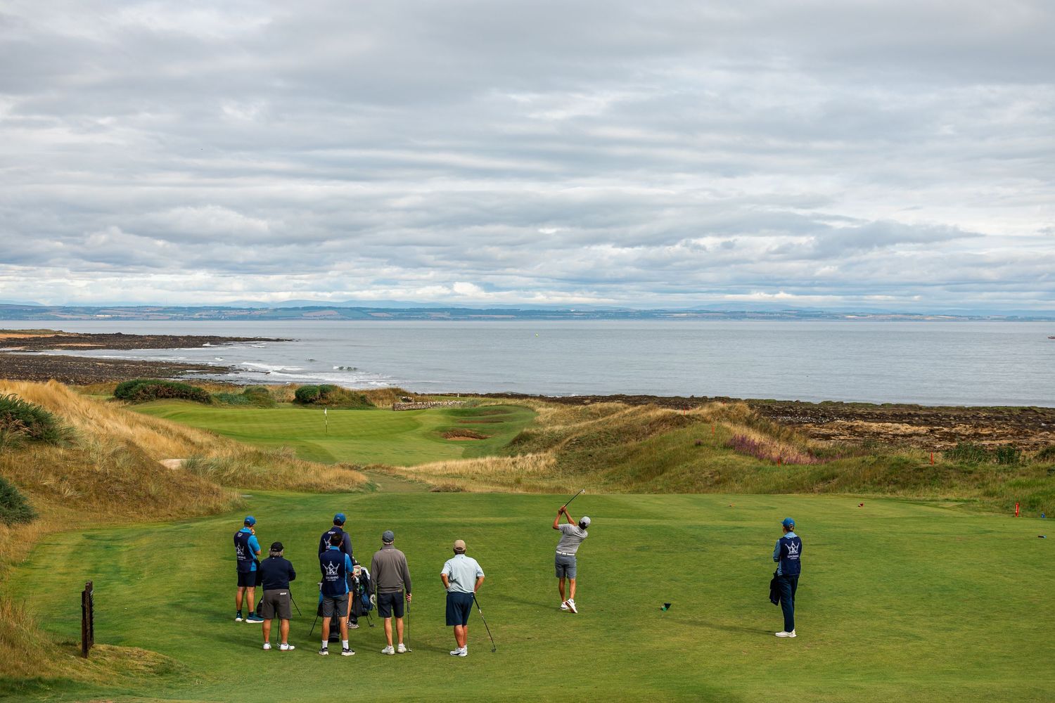 A group of golfers and caddies observe a shot on a stunning coastal links course with ocean views.
