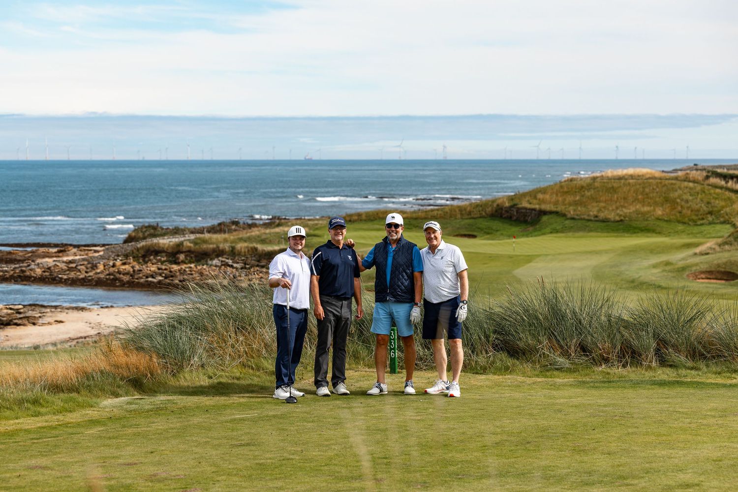 Four golfers pose together on a scenic coastal golf course with ocean views and sandy dunes in the background.