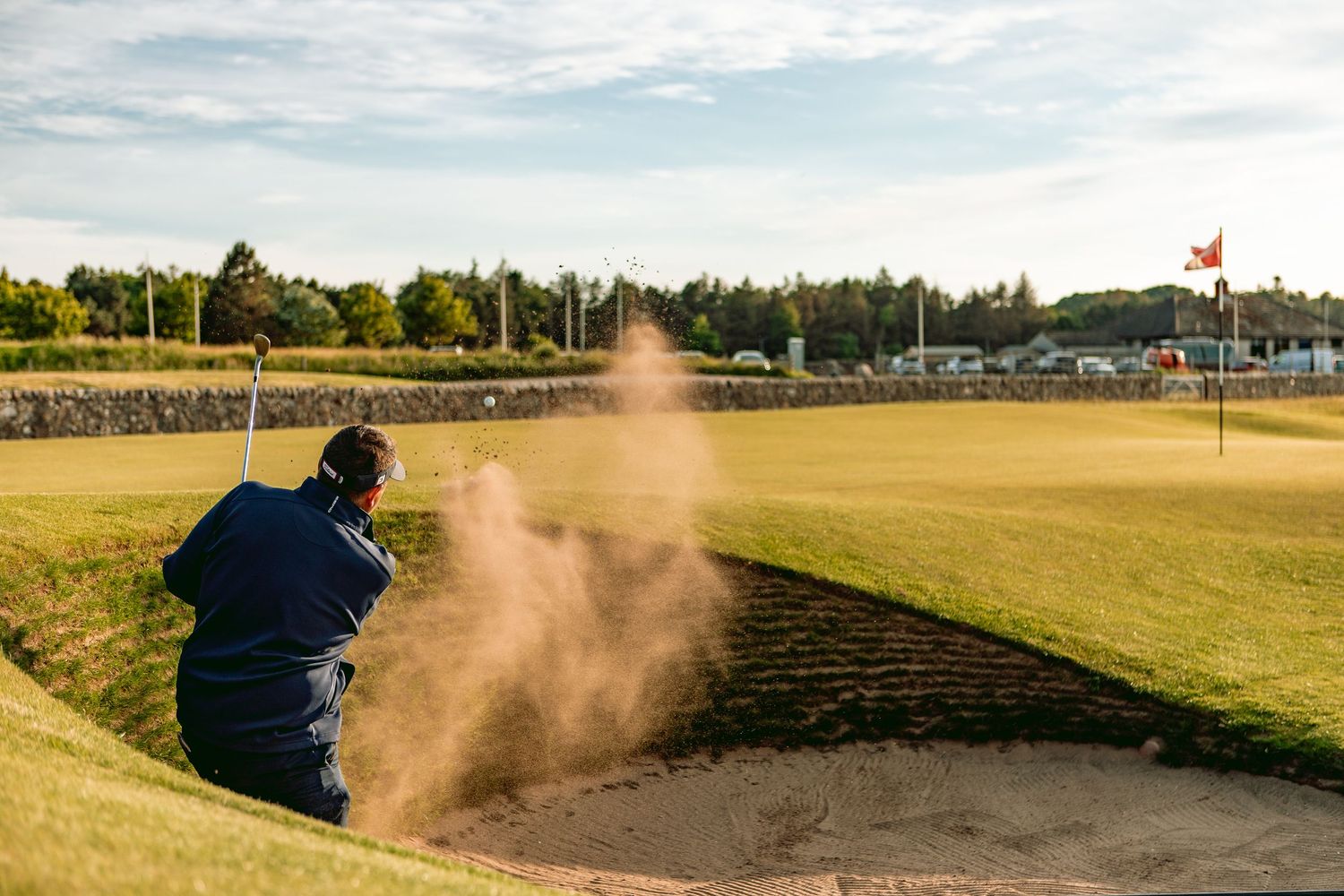 A golfer hits from a sand bunker at Old Course, creating a cloud of dust.