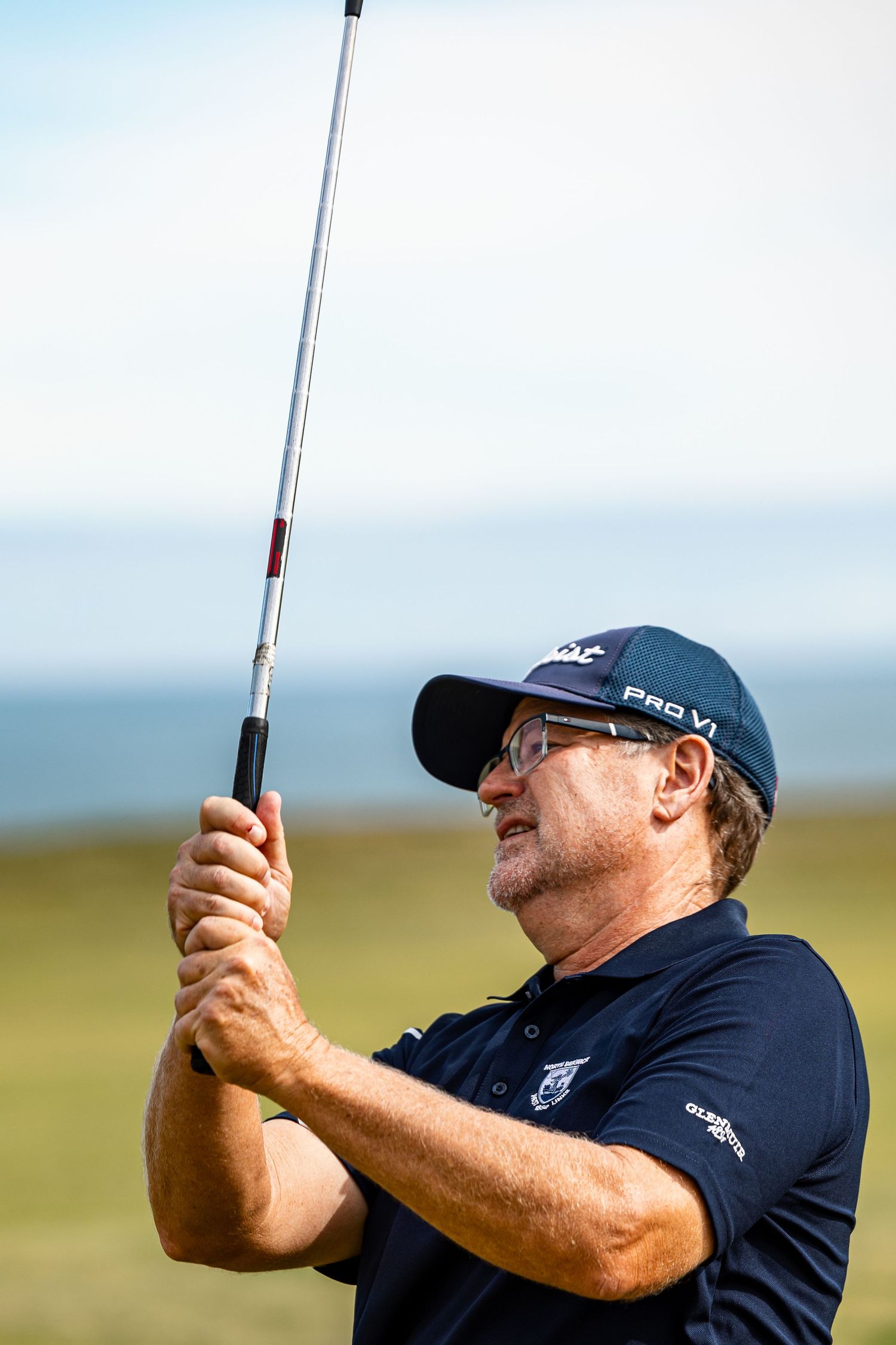 Close-up of a male golfer in a navy polo and Pro V1 cap watching his shot on a seaside golf course.