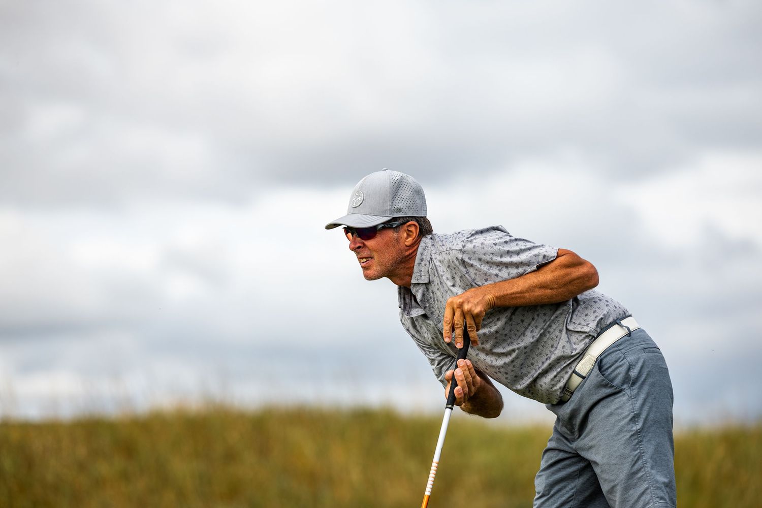 A focused golfer crouches low to read a putt on a windswept links course under cloudy skies.