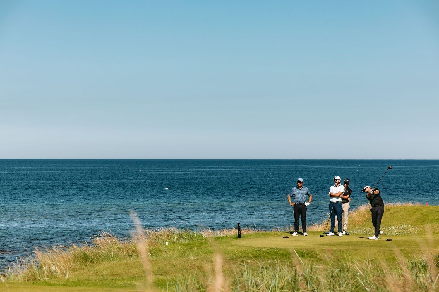 A group of golfers standing at Kingsbarns Links with one golfer in action, playing shot.