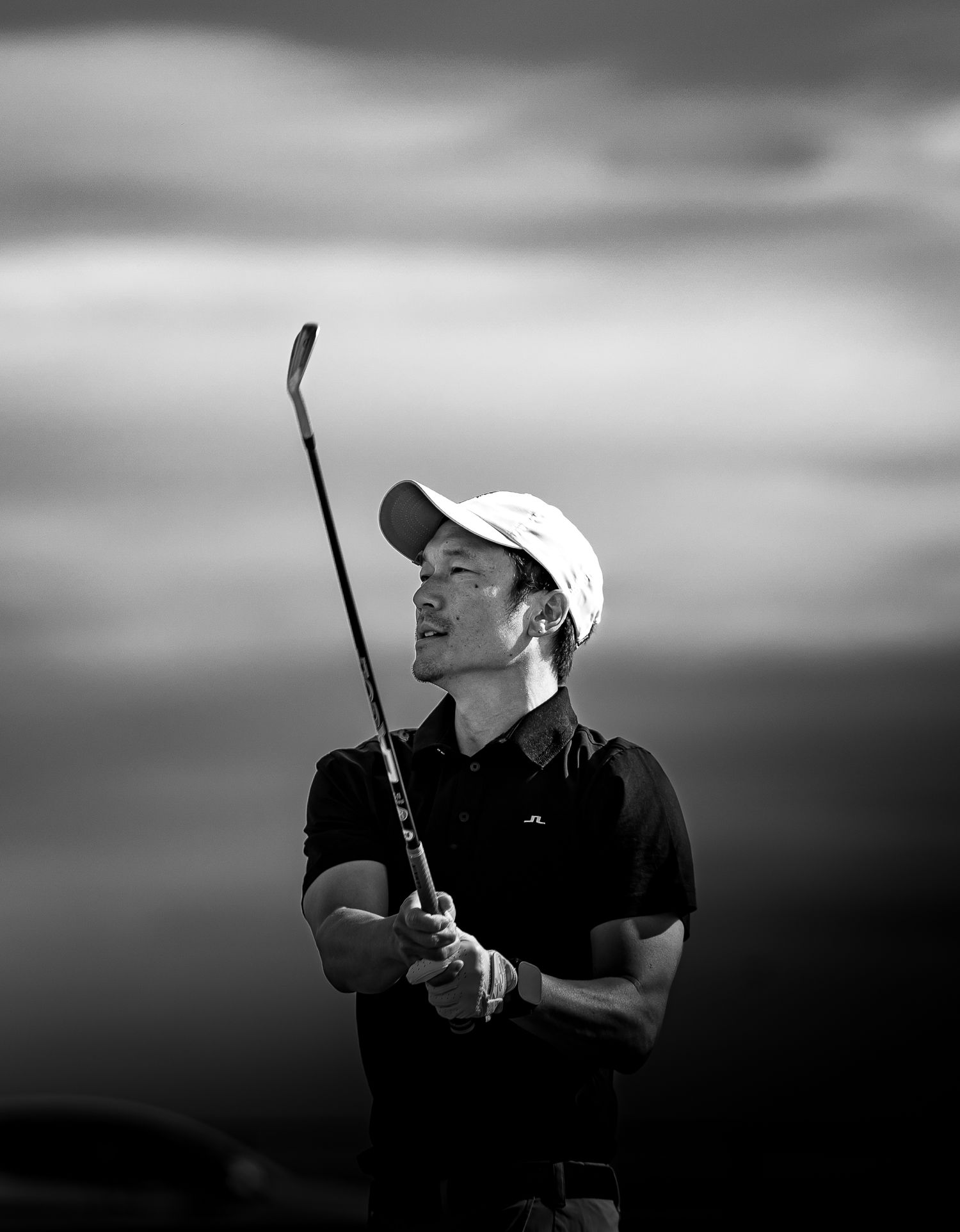 Man wearing white cap with golf club in hand, in action after playing shot.