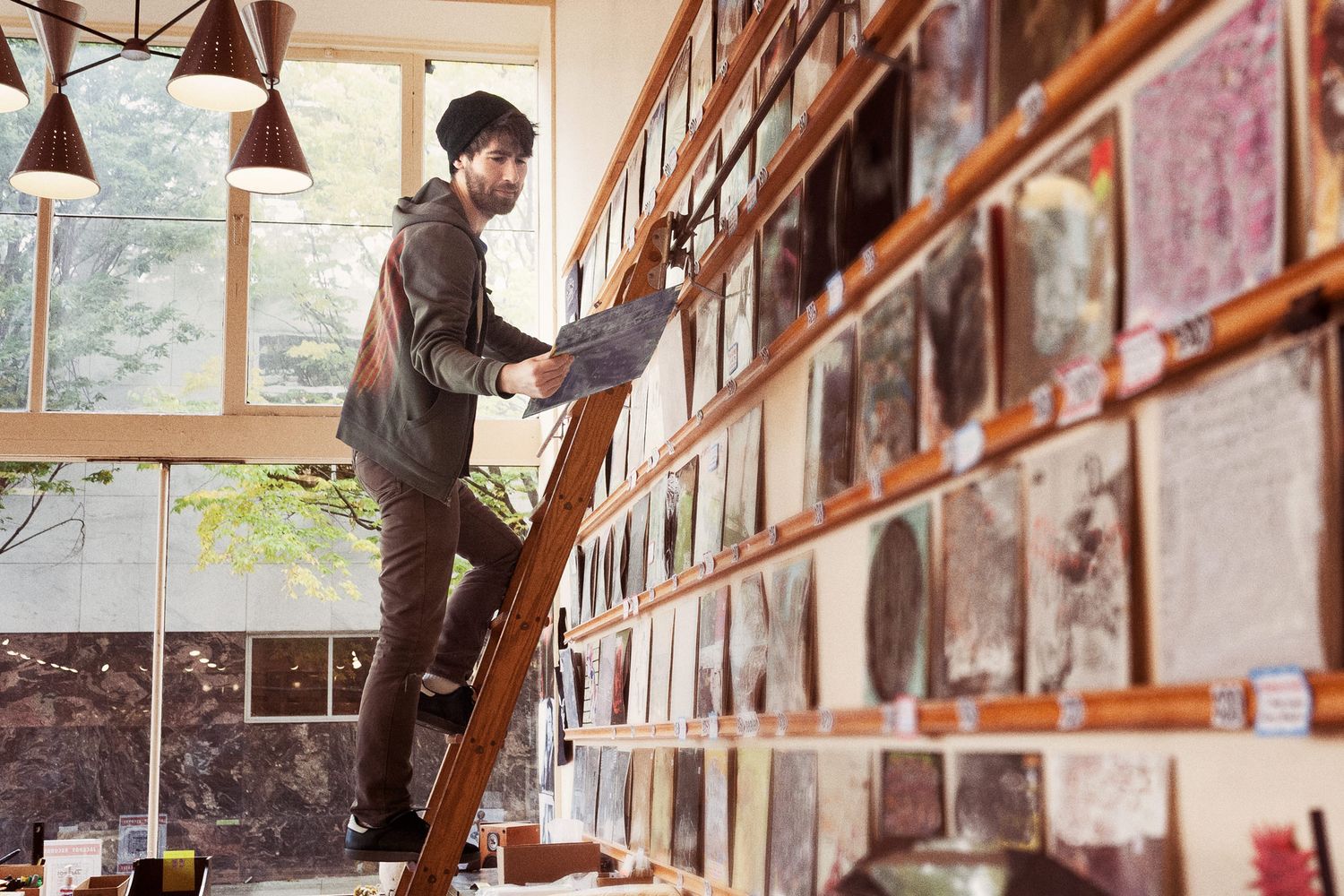 Vinyls line the wall as an employee at a record shop in Portland stands on a ladder holding an album as they read the back of the cover.