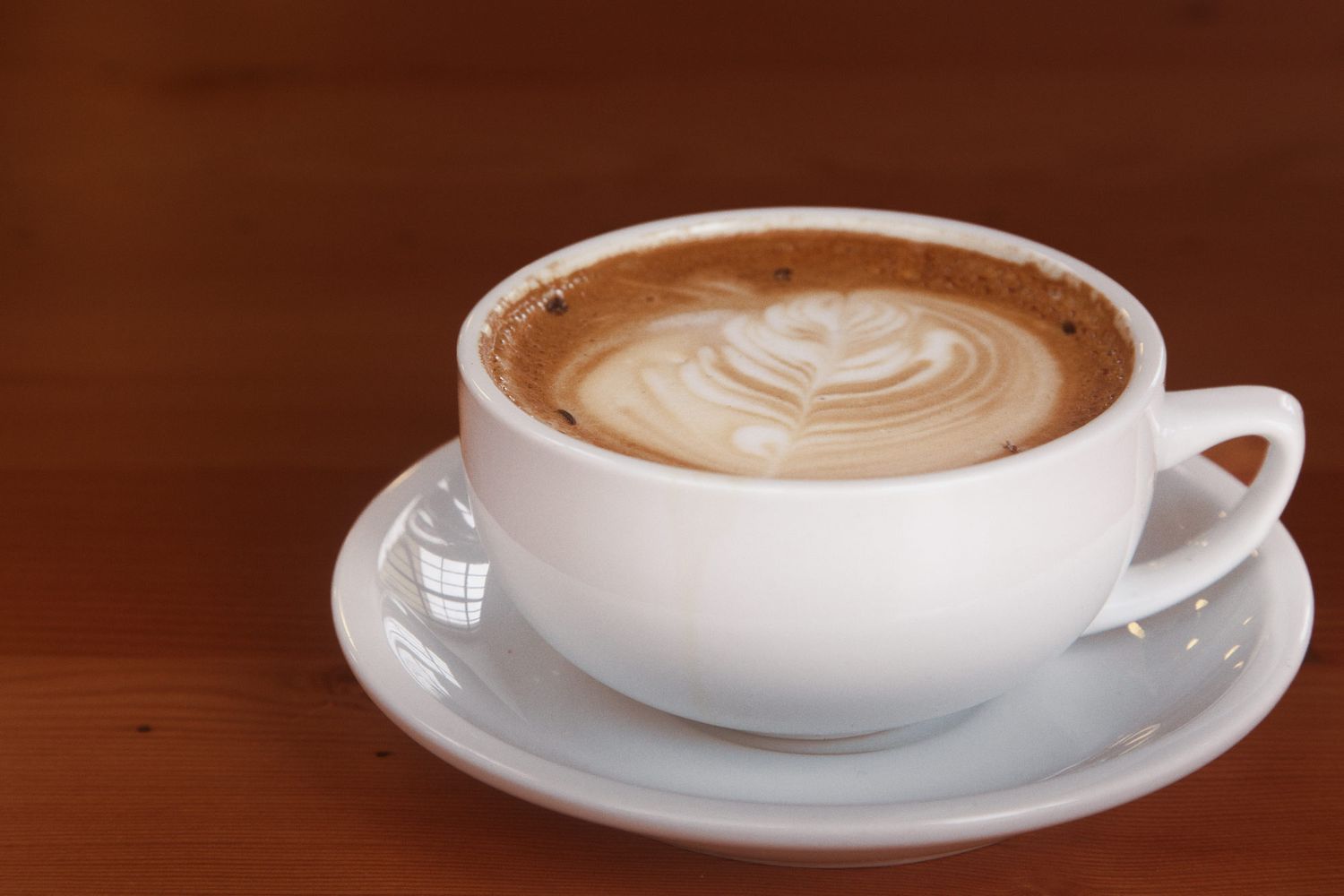 A cup of latte coffee with intricate heart-shaped latte art design on a white saucer against a dark wooden surface.