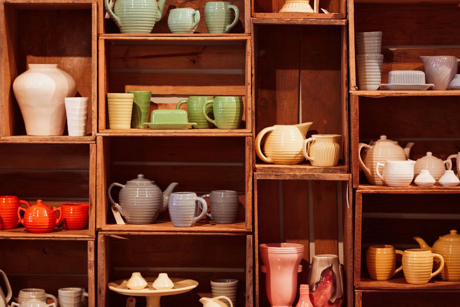Wooden boxes arranged as shelves hold various colored ceramics of teapots, mugs and dishware.