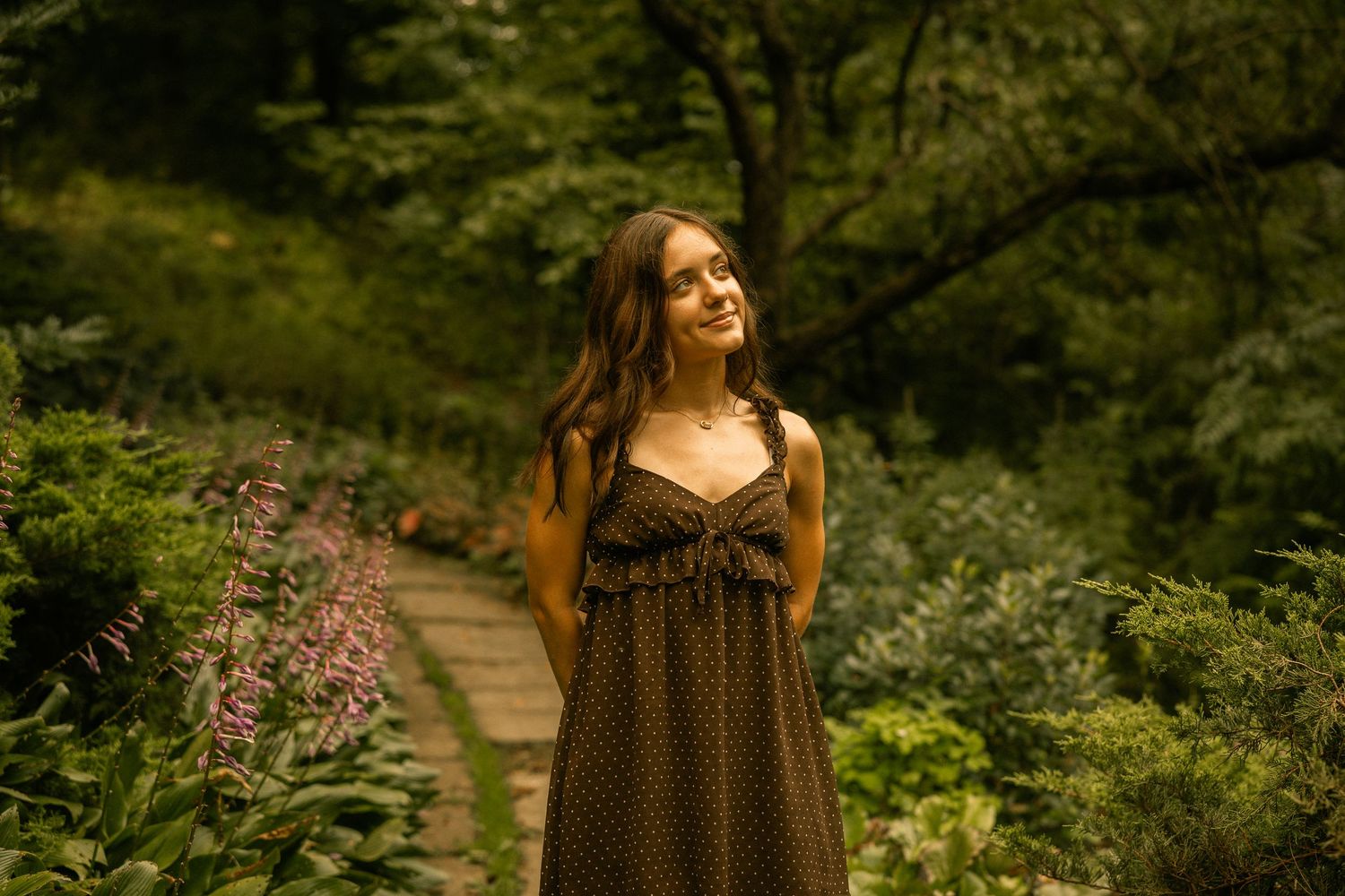 Young woman in olive dotted sundress smiling among lush garden greenery on a stone path.