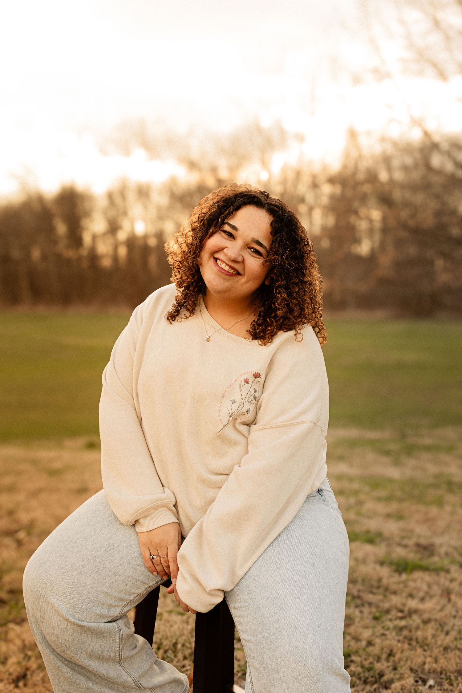 Person in light sweater and jeans smiling during sunset photoshoot in grassy field.