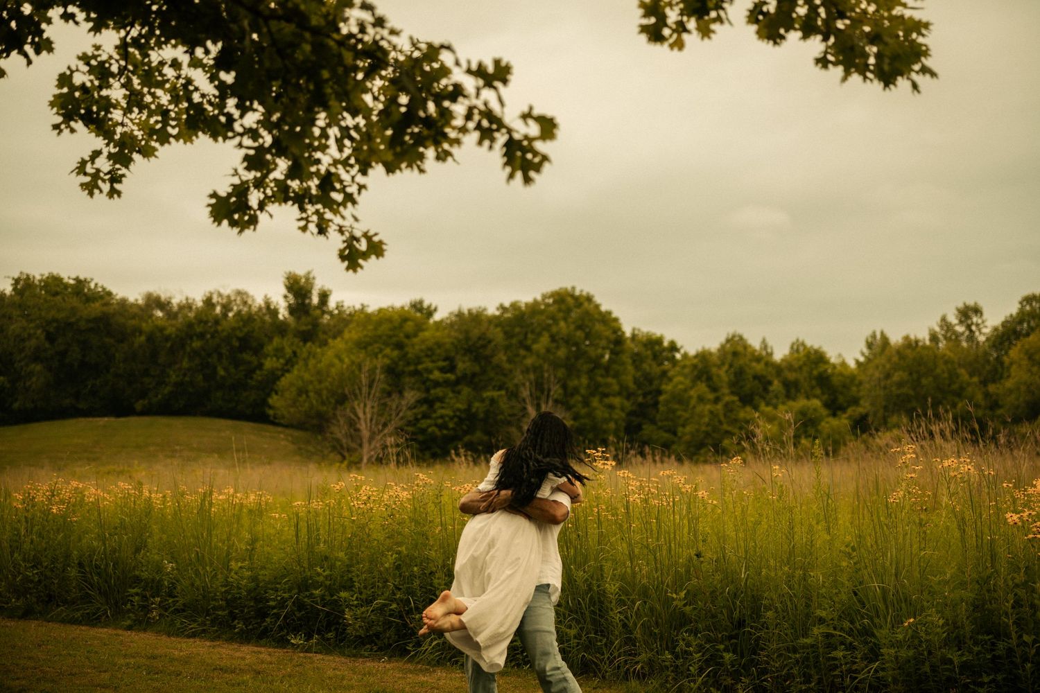 Couple sharing a piggyback ride in a lush green meadow surrounded by trees on a cloudy day.
