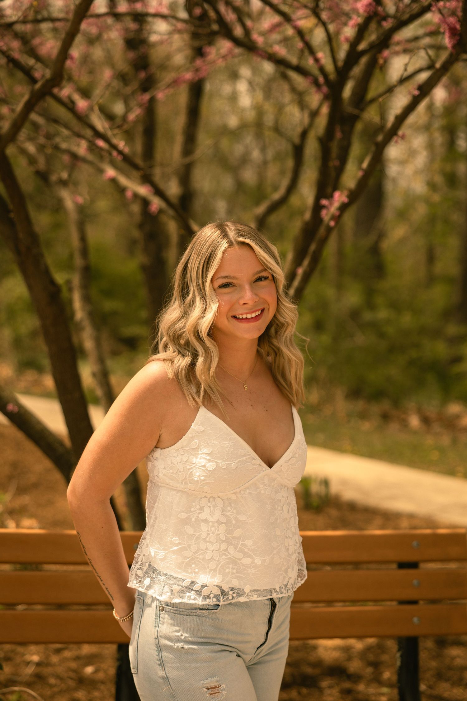 Smiling blonde woman in white lace top and jeans poses by a wooden bench with blooming pink trees in spring park.