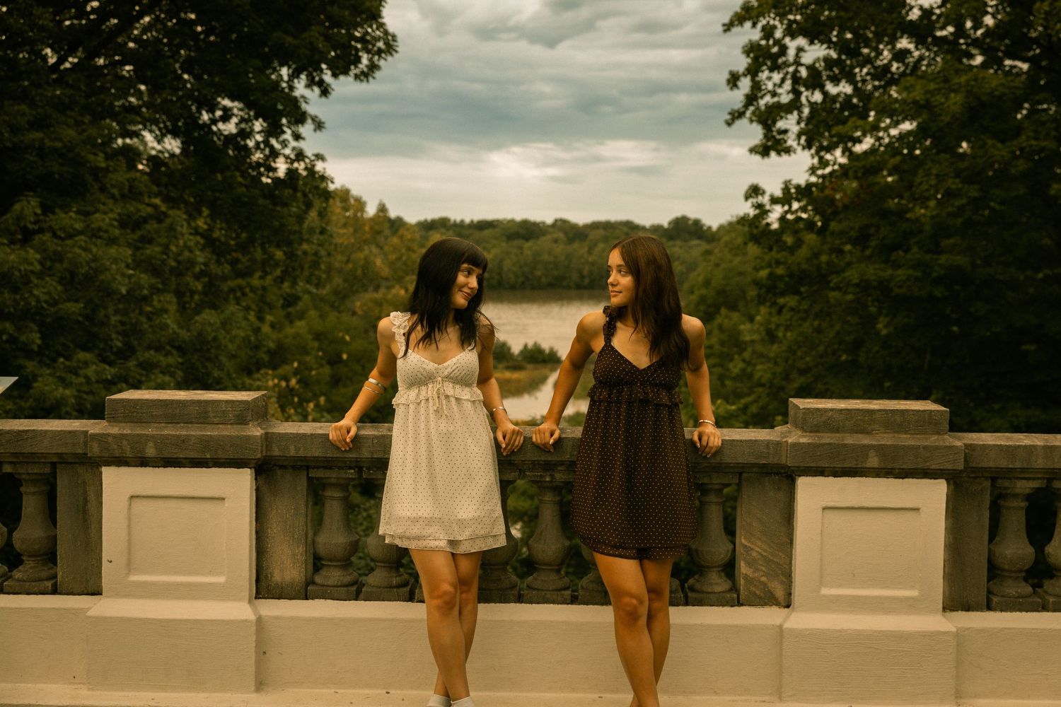 Two women in summer dresses hold hands on a stone terrace overlooking a serene lake surrounded by lush green trees.