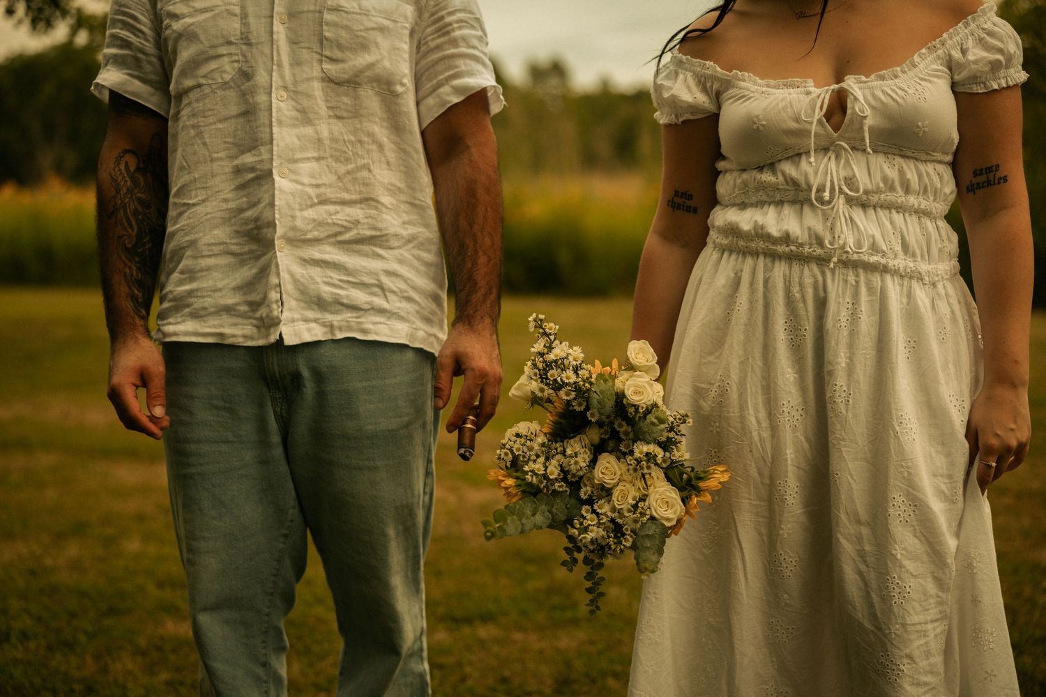 Couple holding hands in a field, bride in white dress with wildflower bouquet, groom in jeans and white shirt.