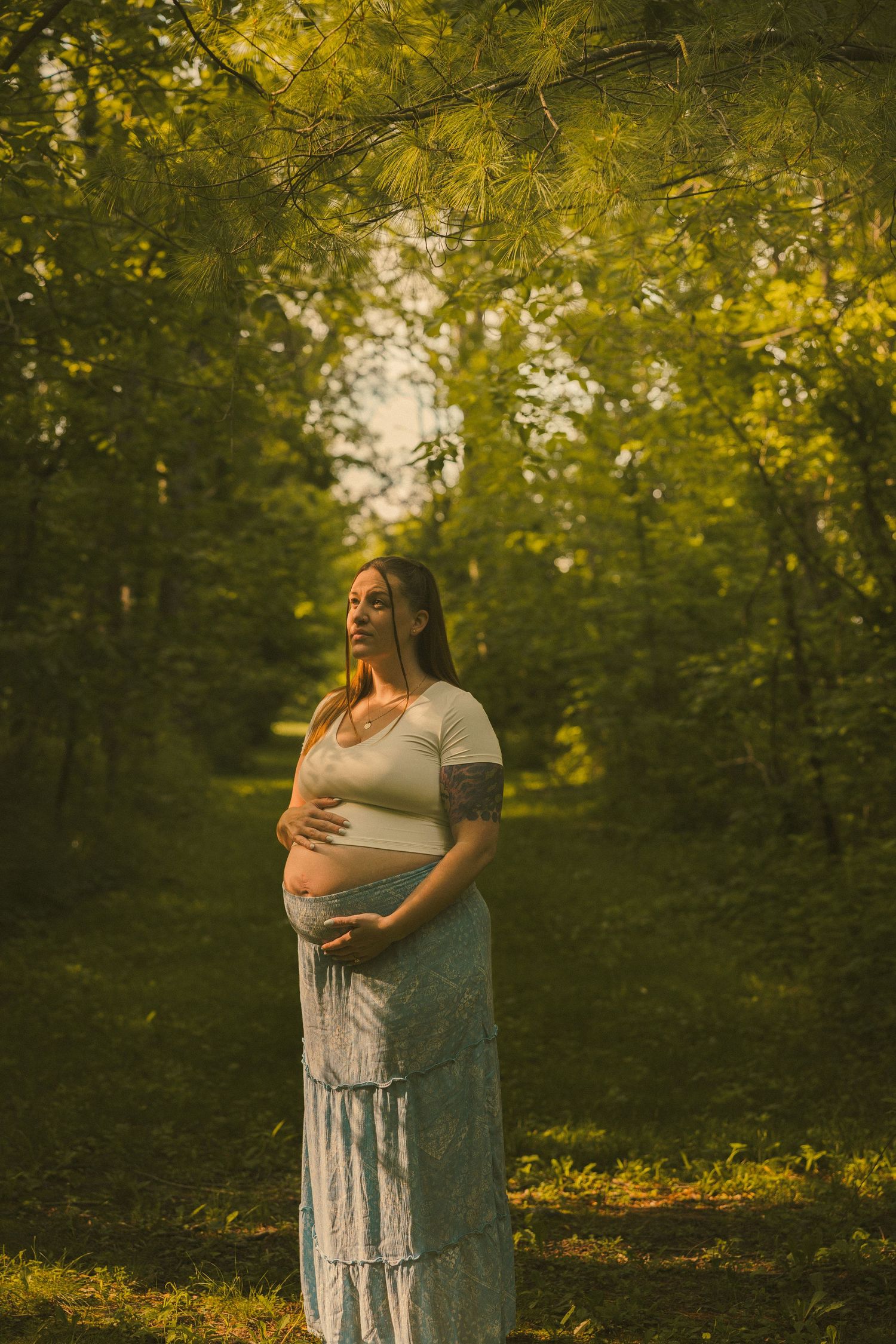 Pregnant woman in white crop top and blue skirt cradling her baby bump outdoors in a sunlit green forest path.
