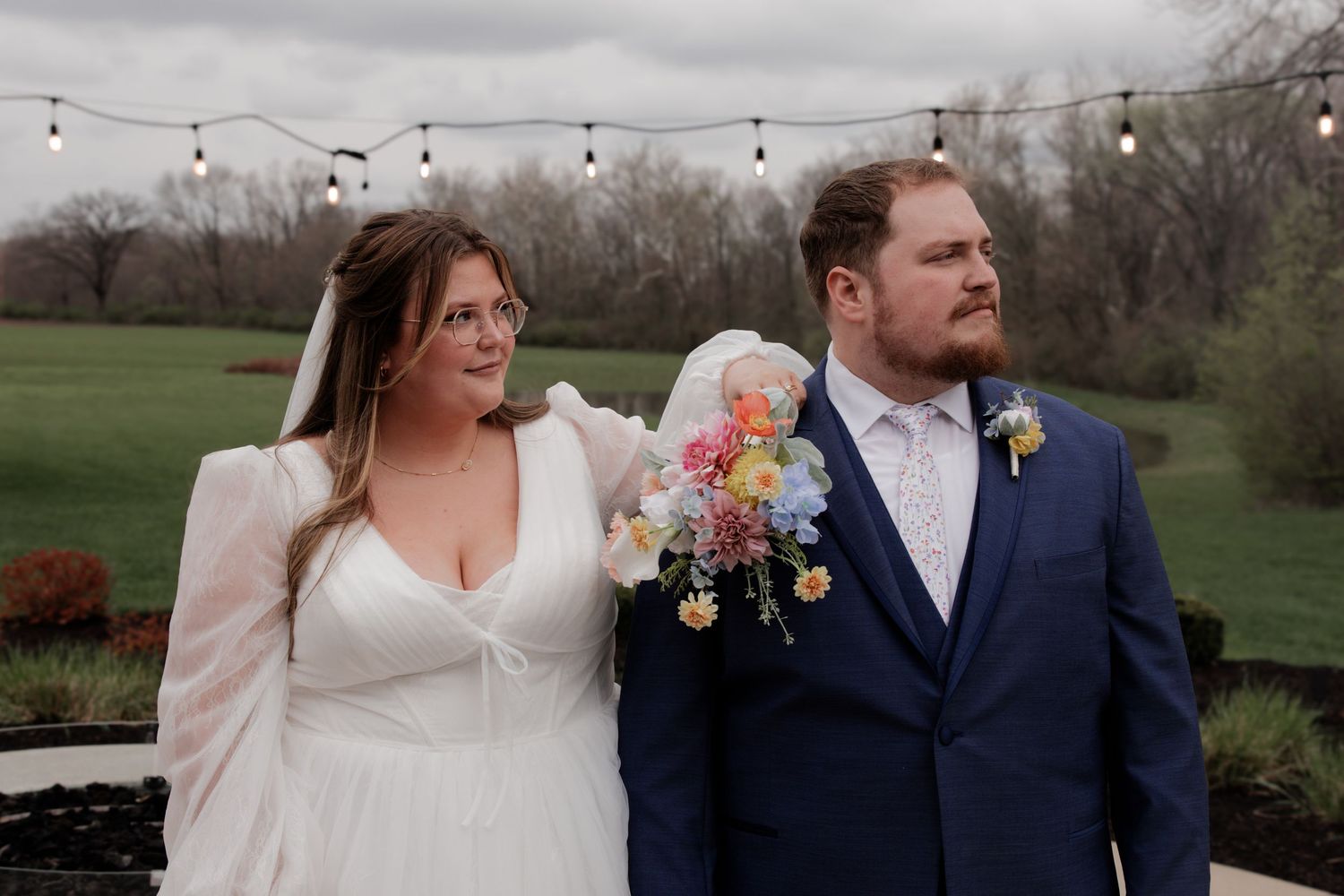 Wedding couple poses outdoors with colorful floral bouquet against natural landscape.