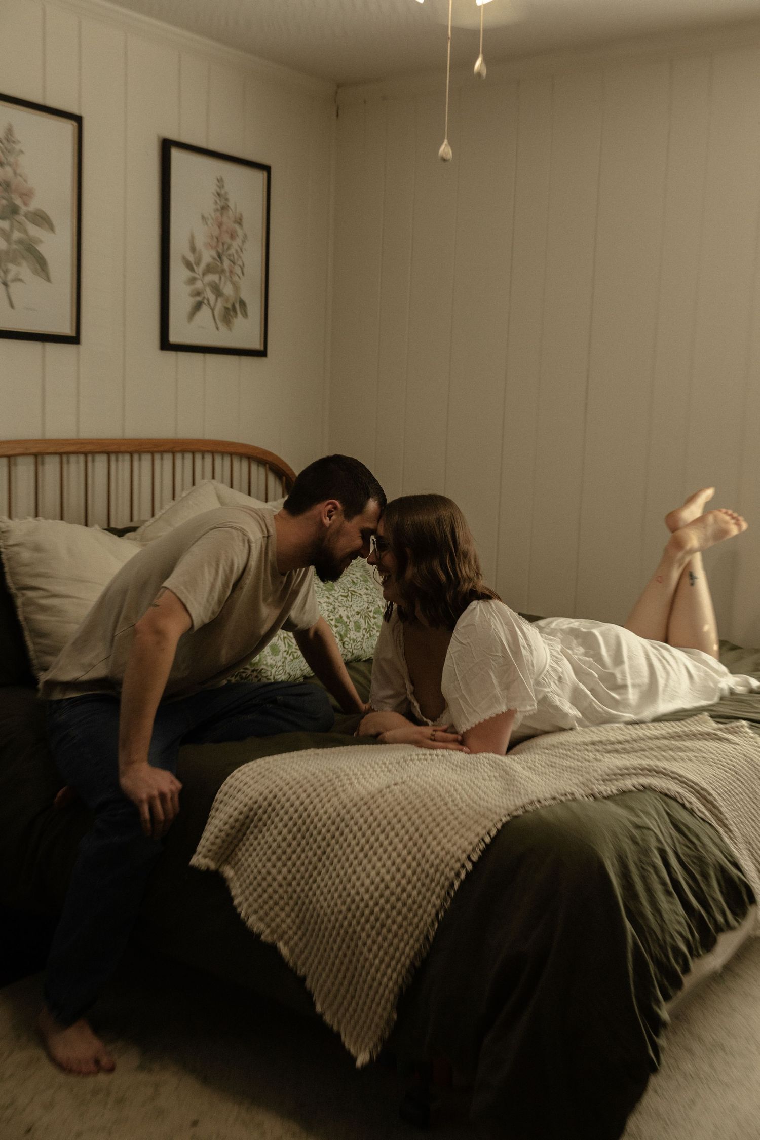 Romantic moment shared between two people on a bed beneath vintage botanical prints and pendant lights.