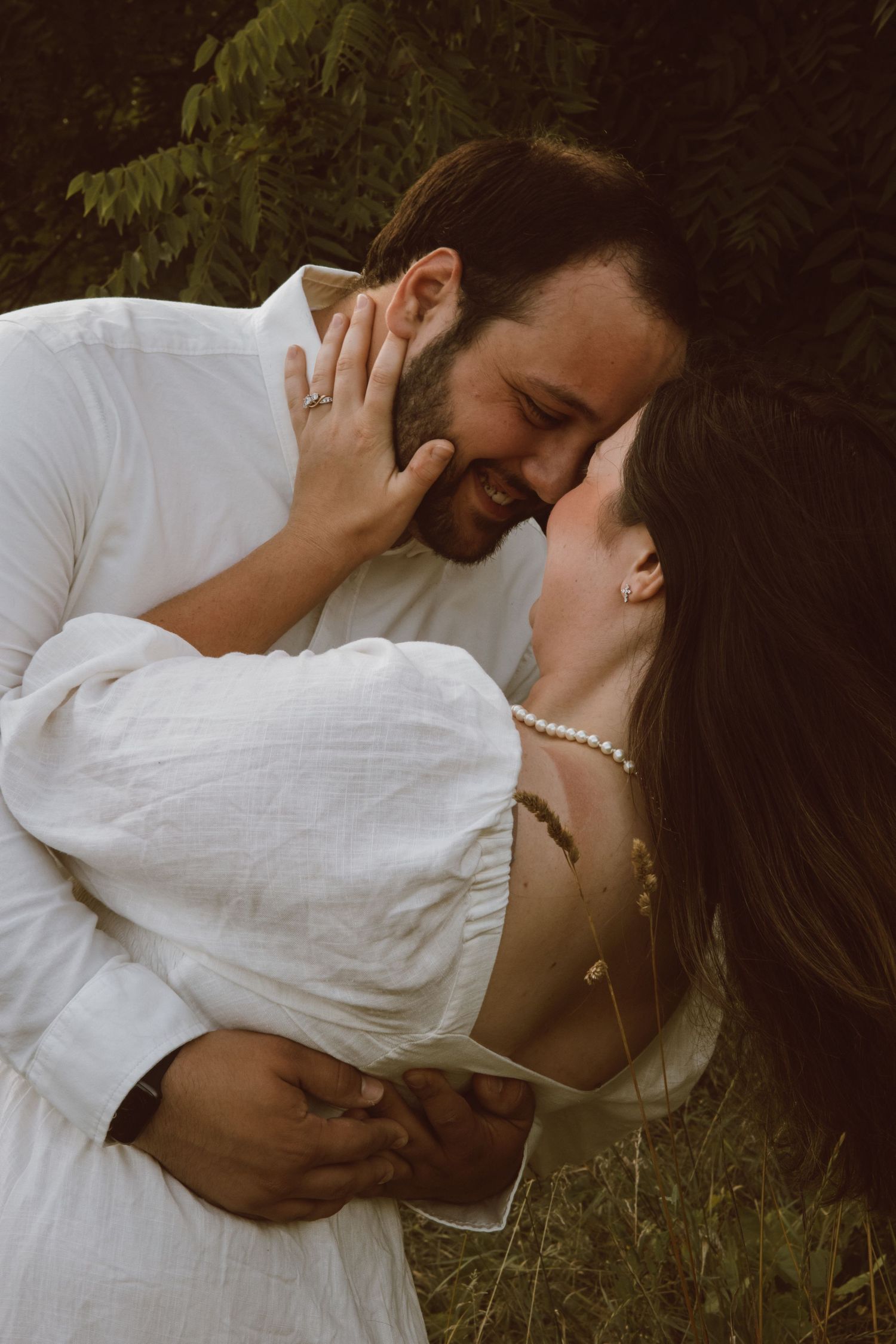 A couple in white clothing shares an intimate embrace in a natural setting.