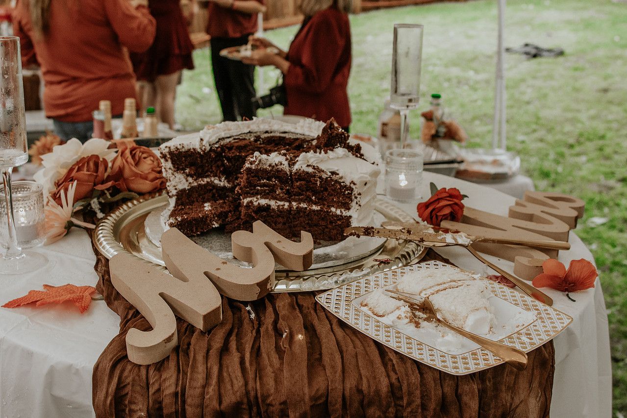 A rustic dessert table displays a layered chocolate cake with white frosting alongside table settings at an outdoor event.