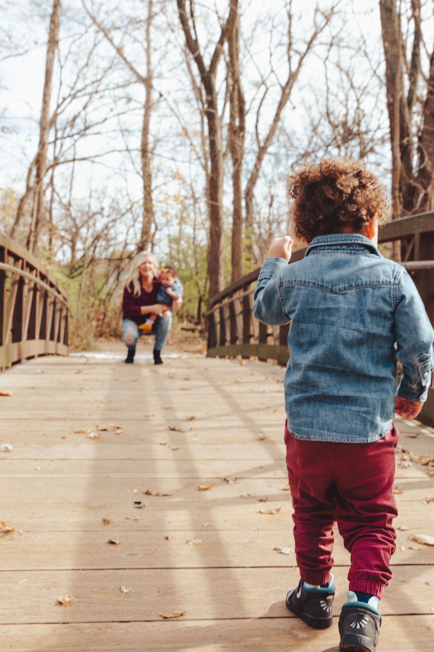Small child in denim jacket and burgundy pants crawling on wooden boardwalk in autumn.