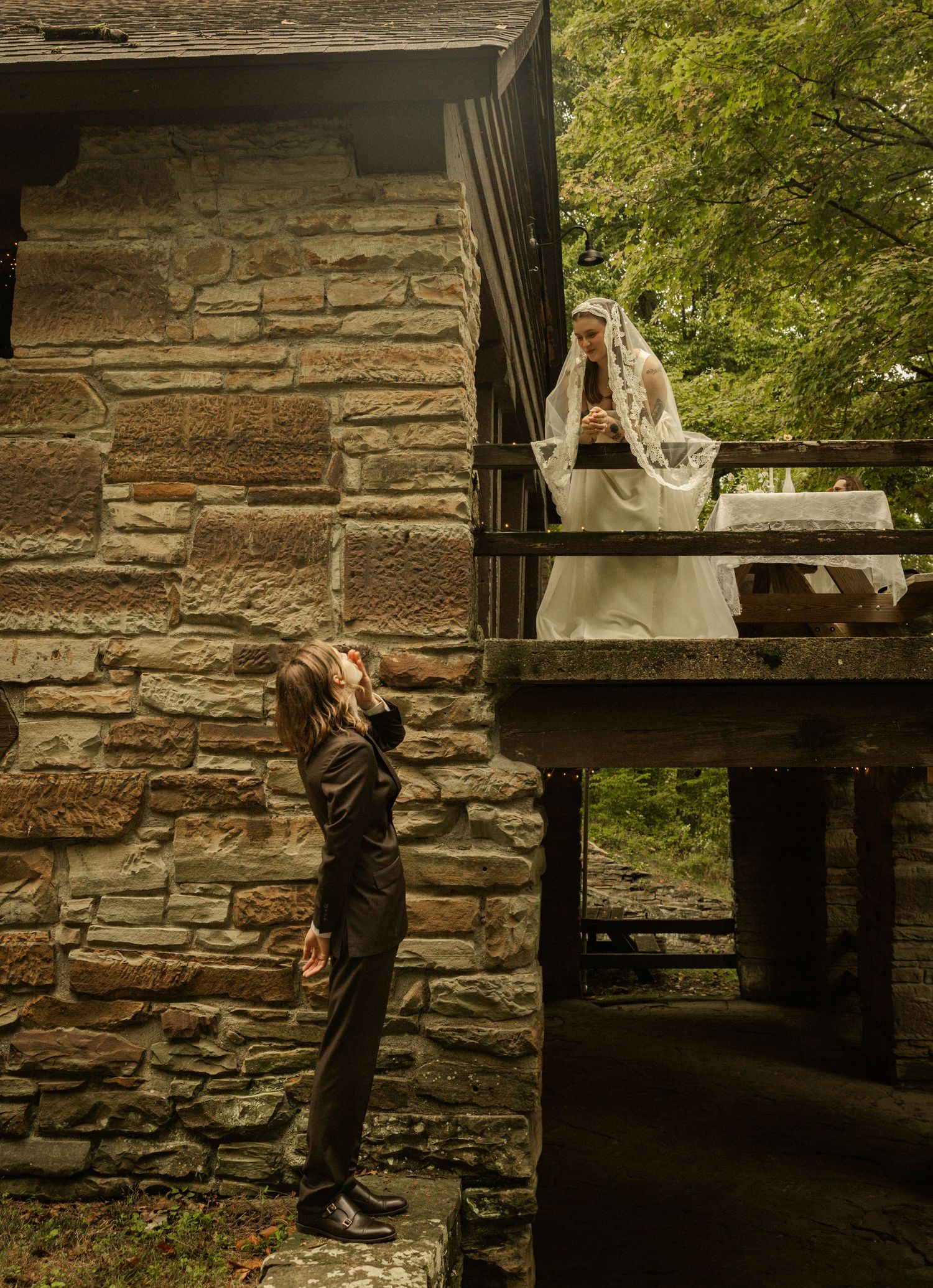 Groom looks up at bride in white lace veil on rustic stone building balcony surrounded by lush greenery.