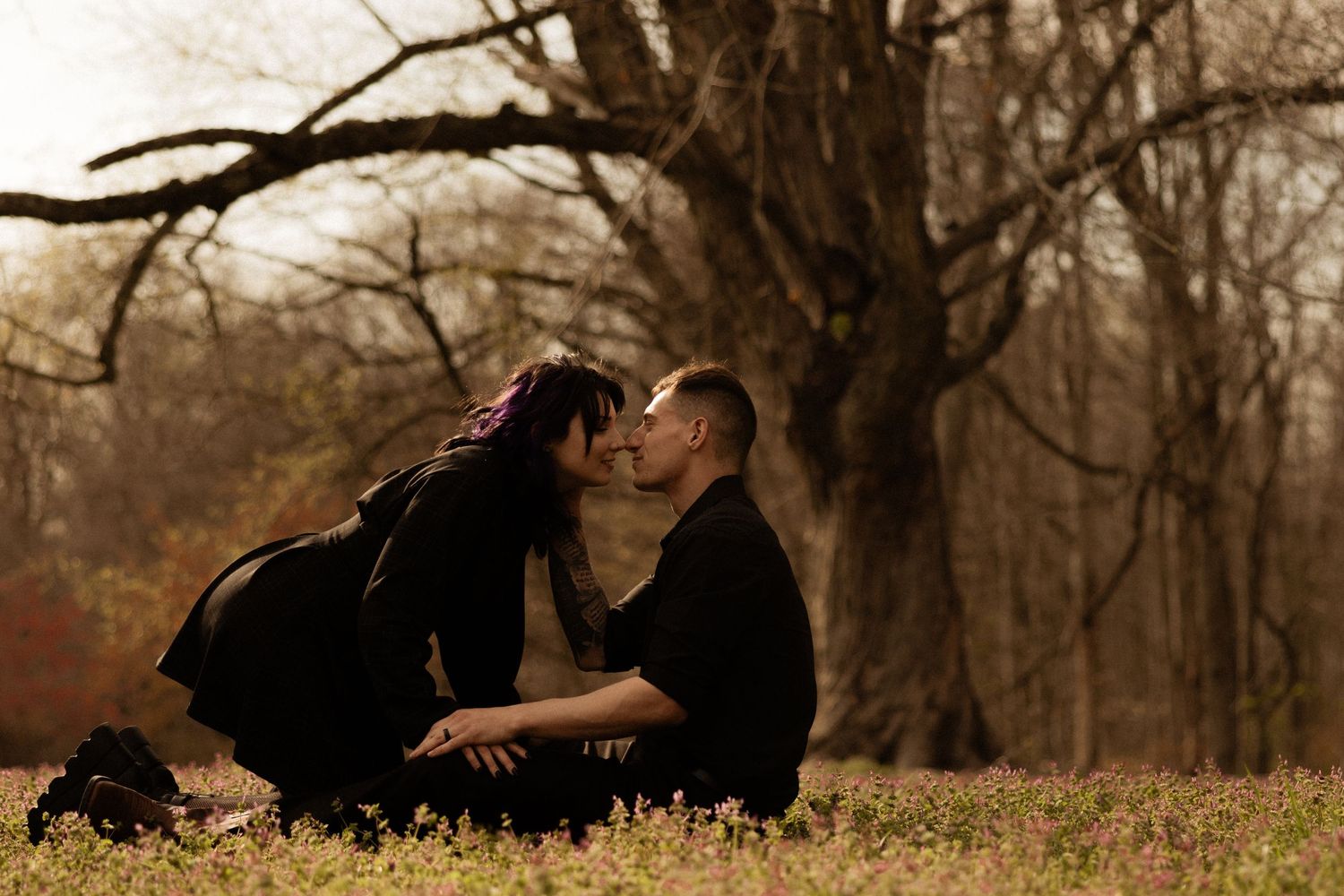 A couple in dark clothing share a romantic moment outdoors with trees in the background.