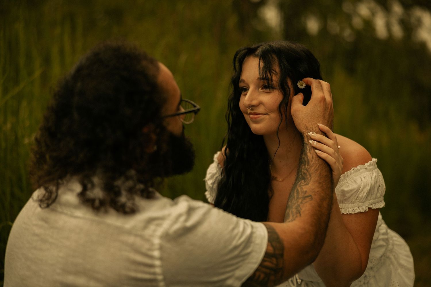 A bearded man with glasses tenderly brushes hair from a smiling tattooed woman in a white dress outdoors.