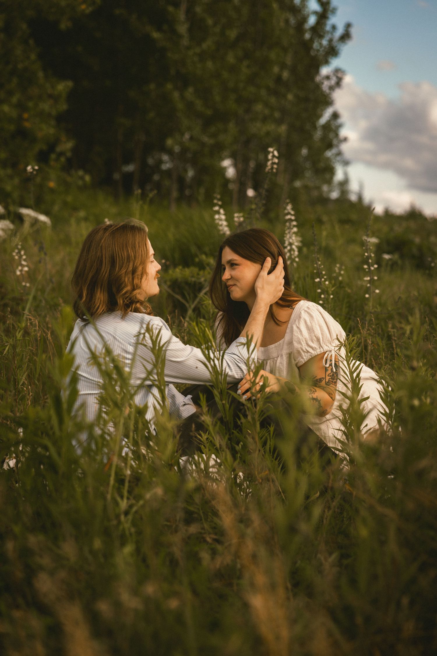 Two women in white dresses sitting together in a lush green meadow, sharing a tender moment at golden hour.