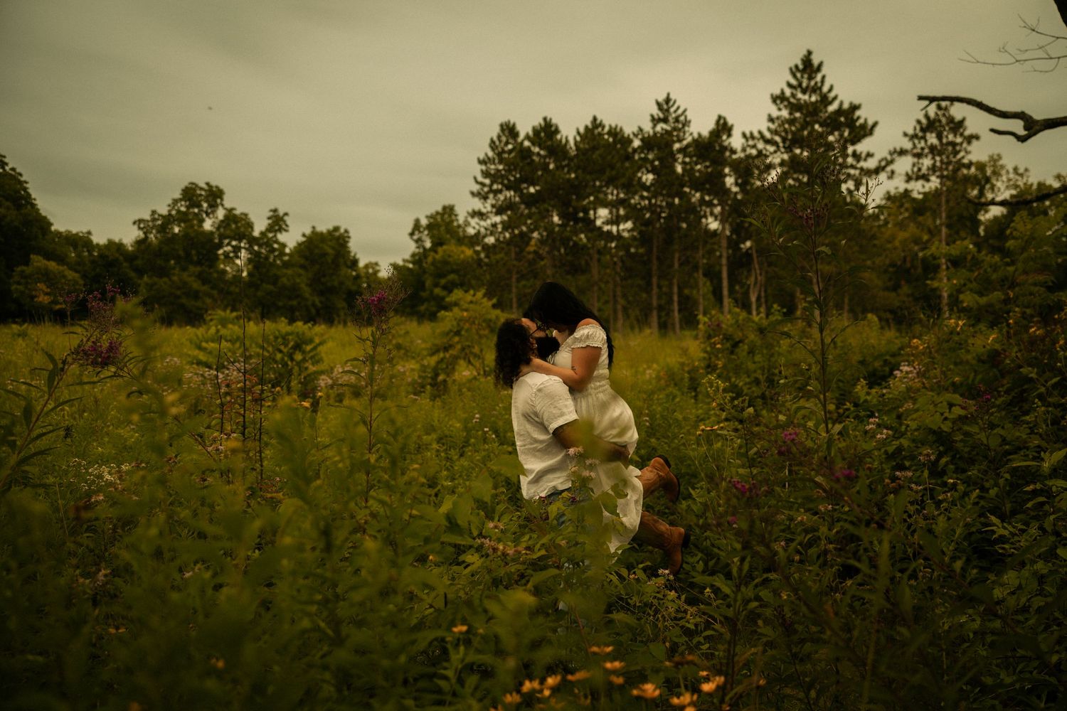 Couple embracing romantically in a lush green meadow surrounded by wildflowers and tall pine trees under overcast skies.
