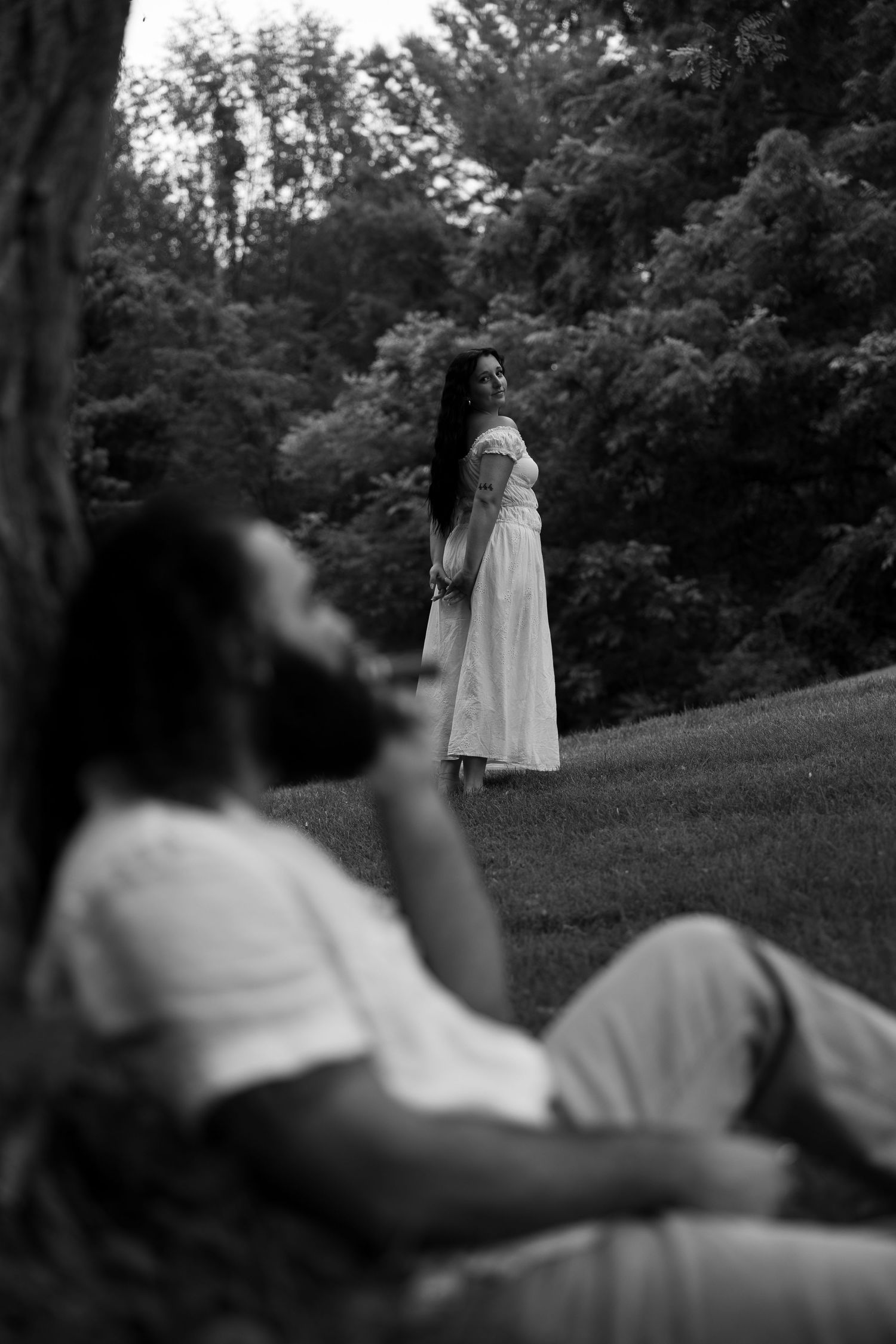 Black and white photo of a couple in a park, woman in white dress standing while man reclines against a tree.