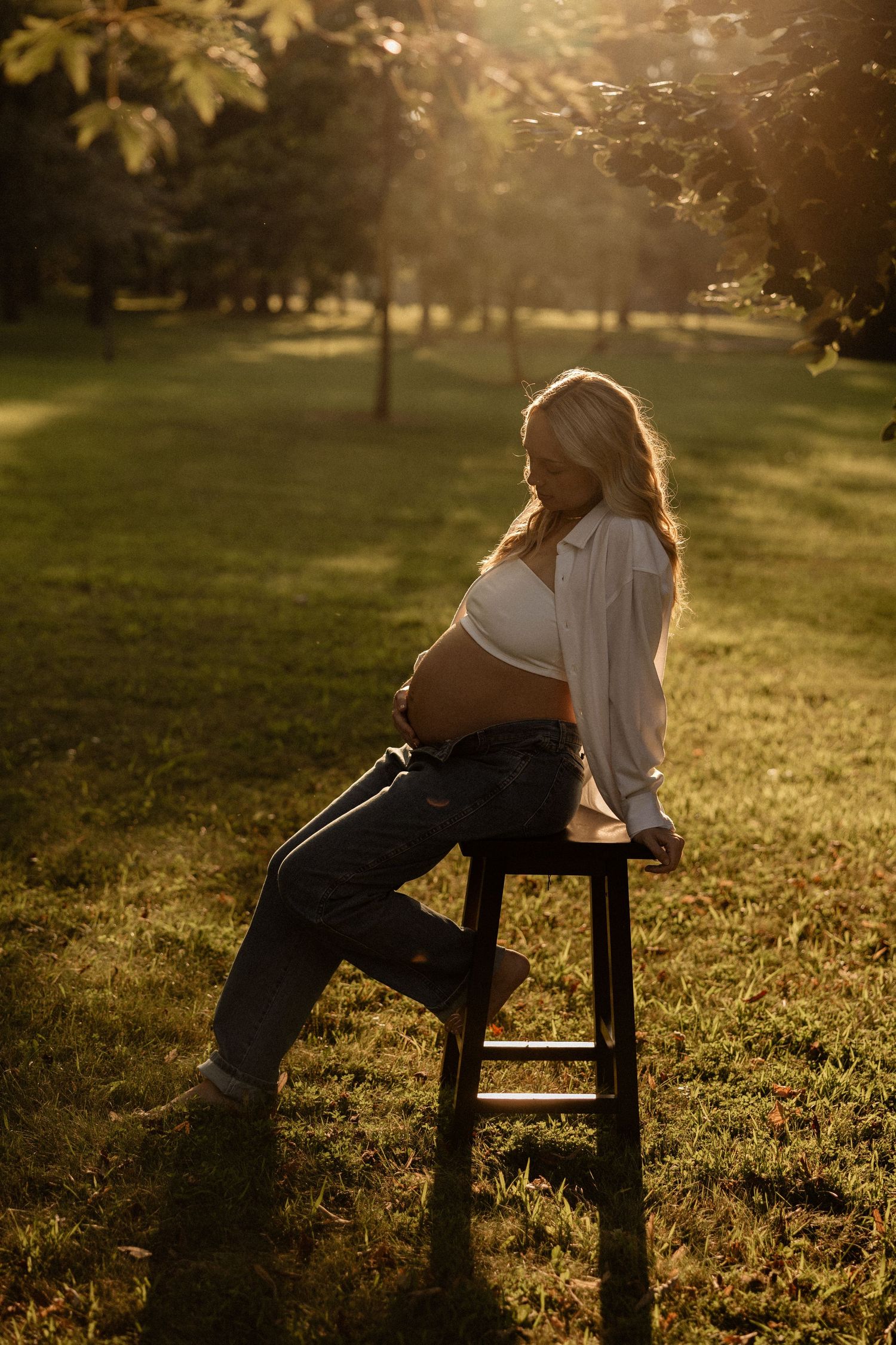 A silhouetted maternity portrait of someone sitting on a stool in a sunlit park.
