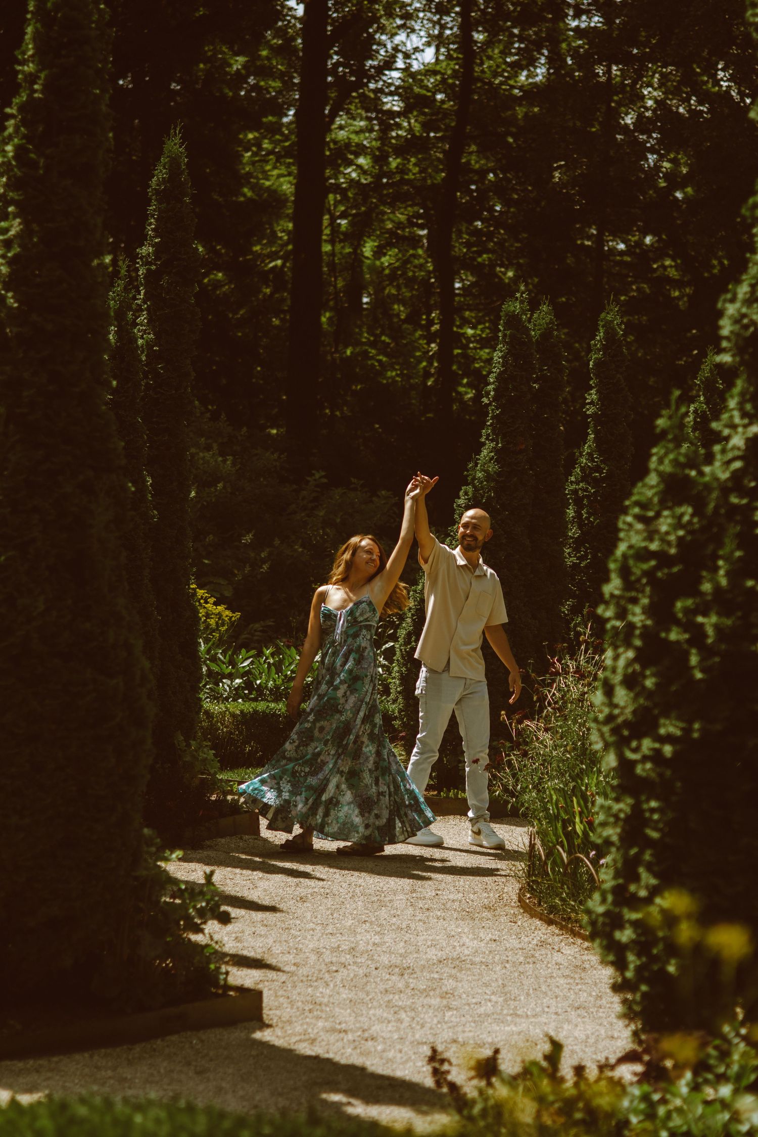 A couple walks hand in hand down a sunlit gravel path through the woods.