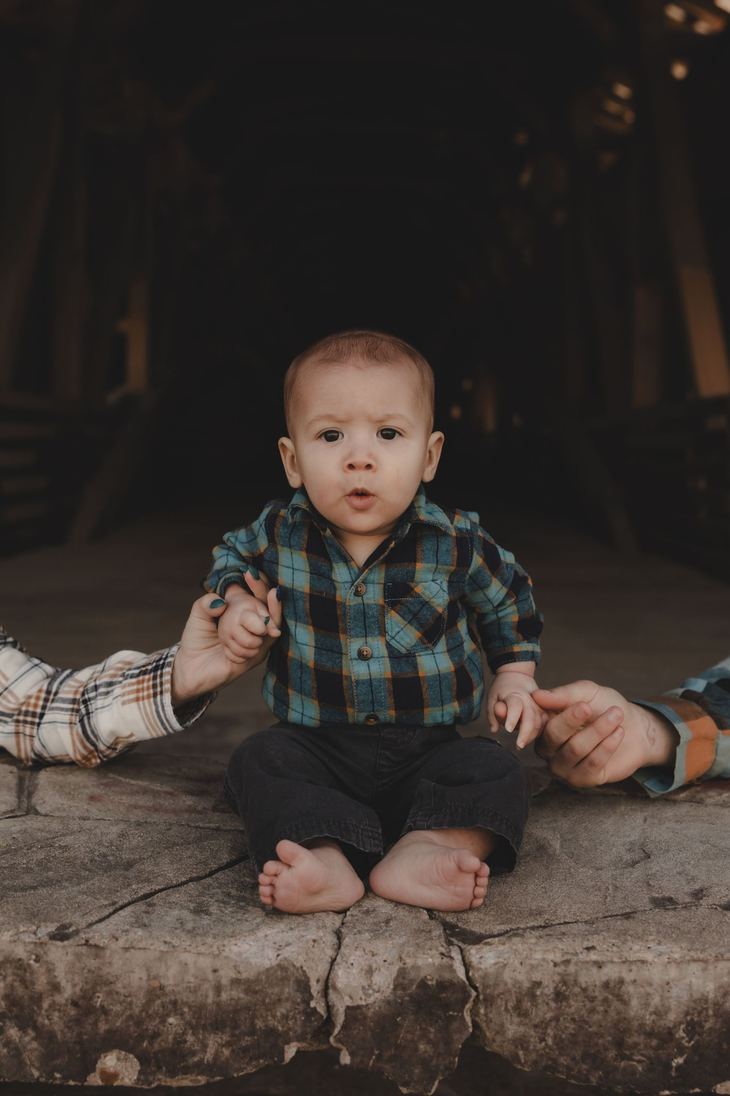Baby in plaid shirt sitting on stone surface during outdoor photoshoot.