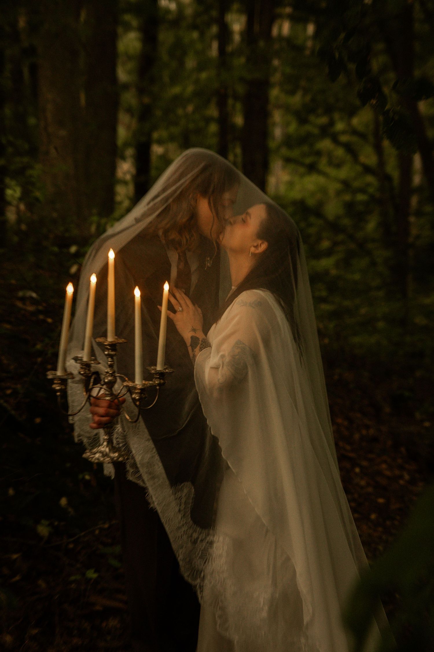 A couple shares a romantic kiss under a veil in a forest, illuminated by candles on an ornate candelabra.