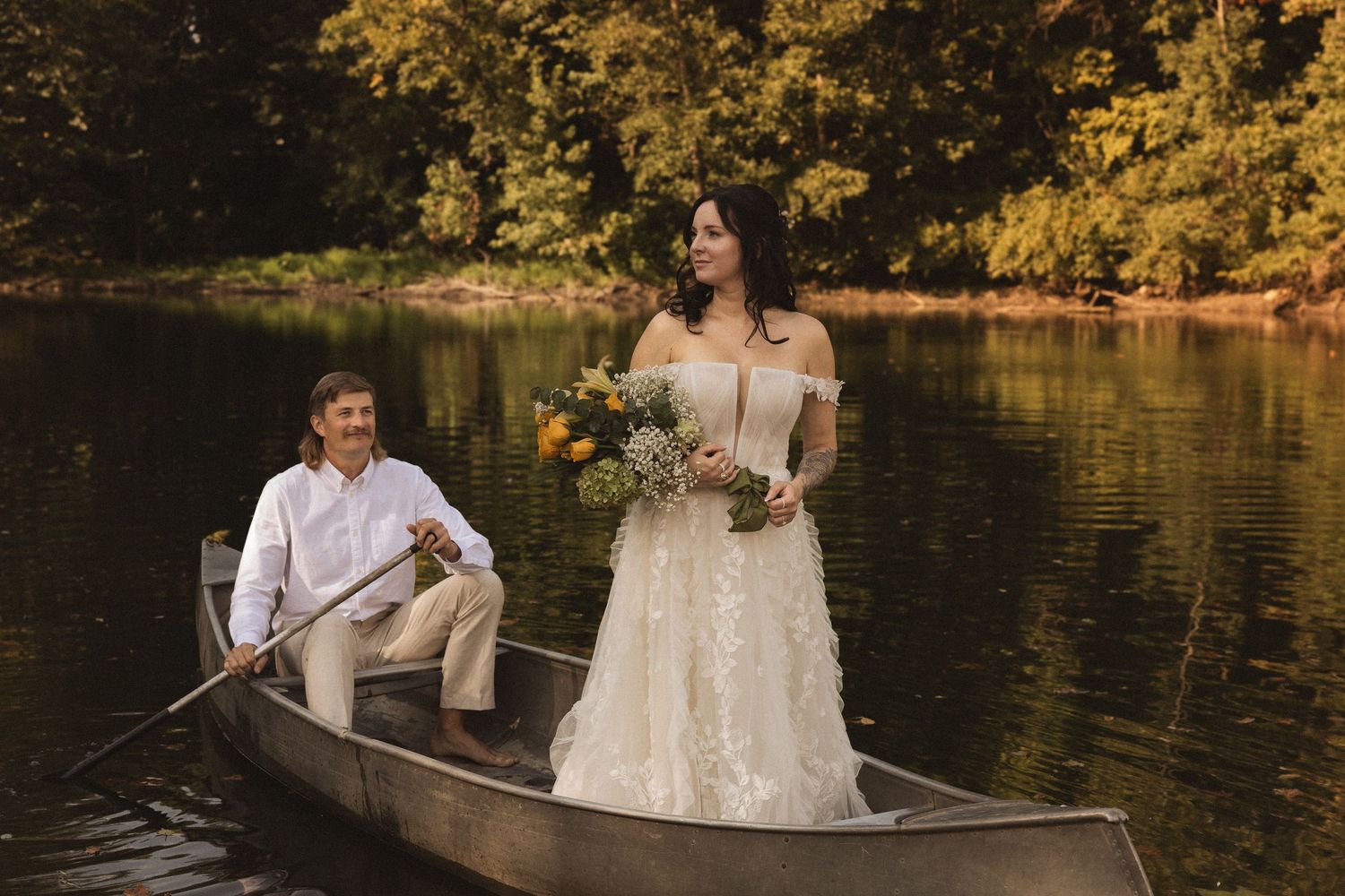 Wedding couple ride in a wooden rowboat across a serene lake surrounded by autumn foliage.