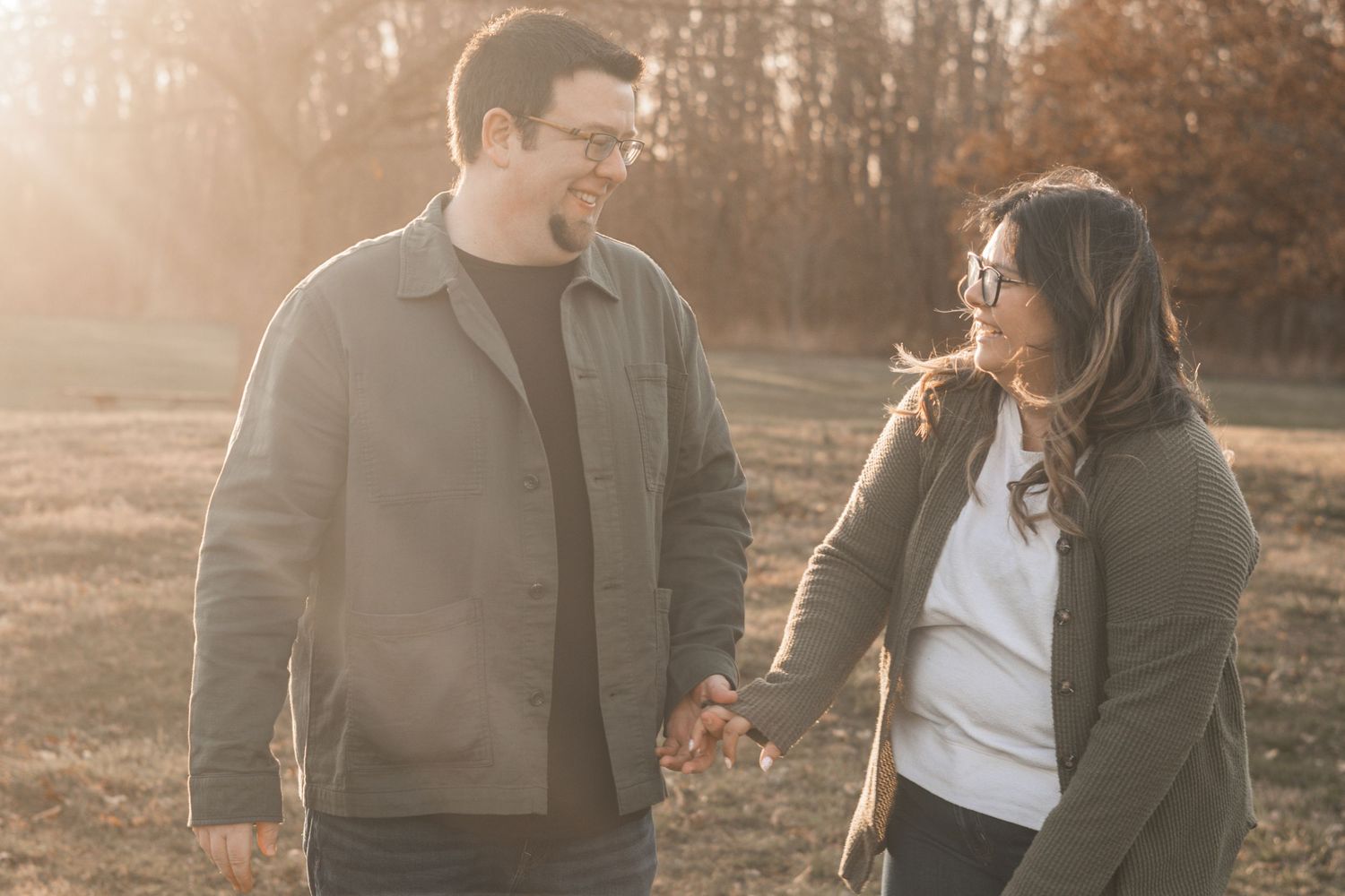 A couple holds hands walking through an autumn forest during golden hour sunset, casting warm glows through the trees.