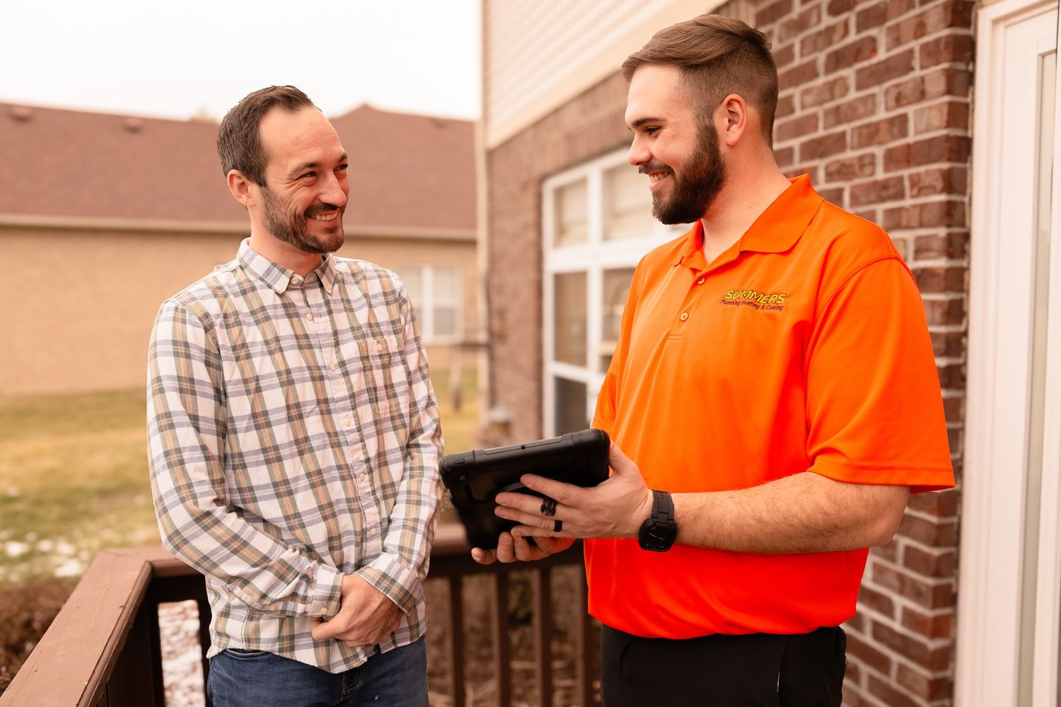 A smiling technician in an orange polo shirt shows a tablet to a homeowner at the front door of a brick house.