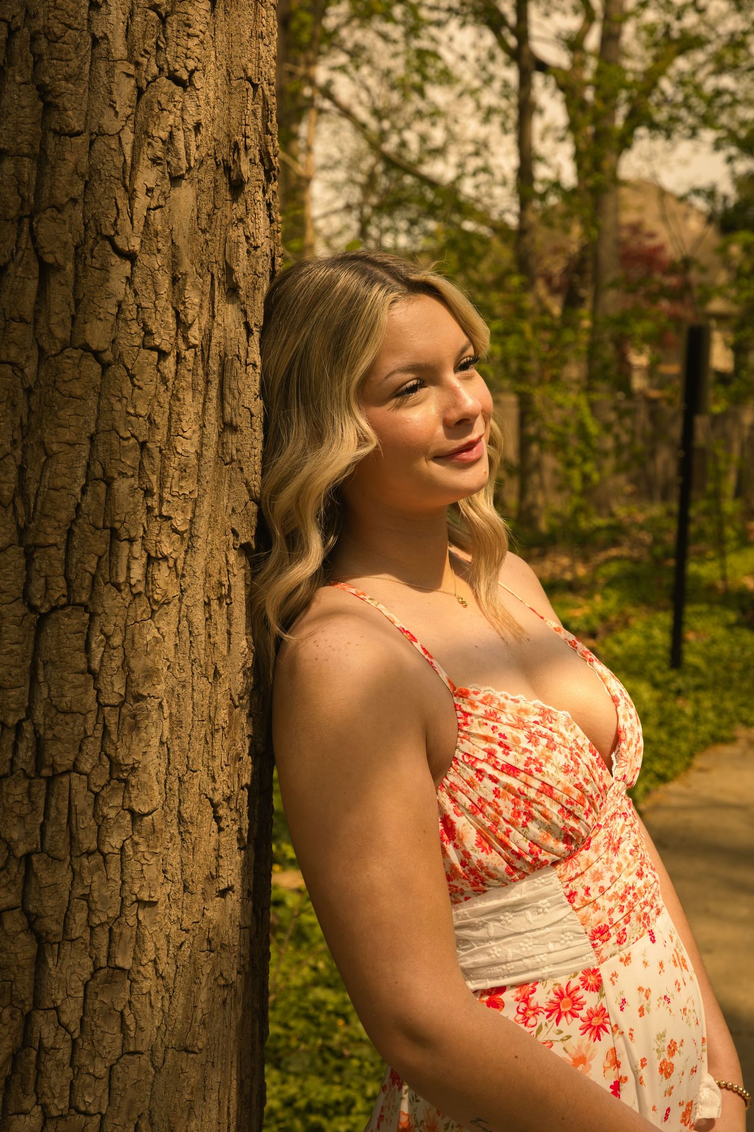 Pregnant blonde woman in floral sundress leaning against tree trunk in park, smiling softly in warm sunlight.
