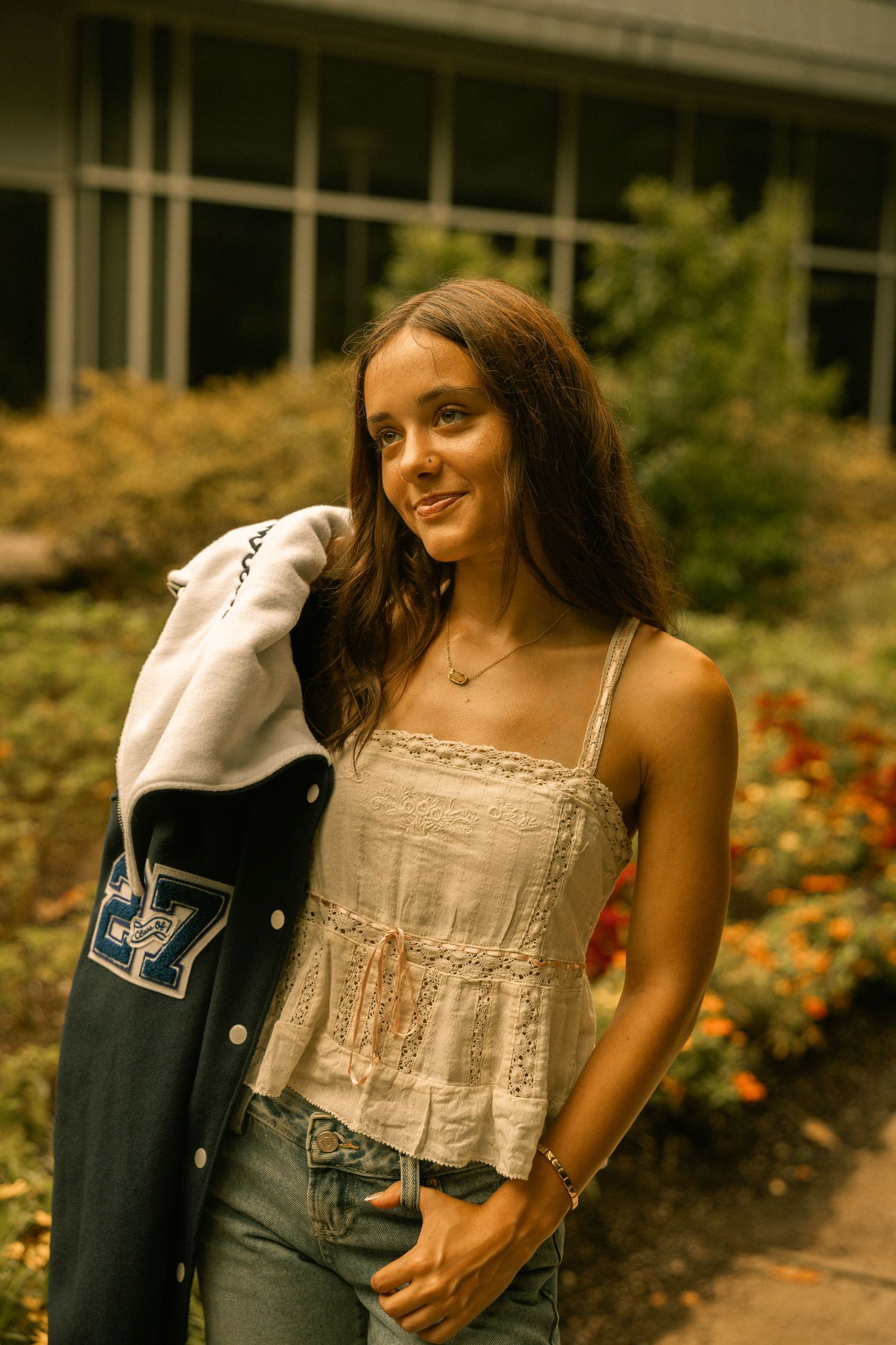 Young woman in white lace top and jeans holding a varsity jacket, smiling outdoors near colorful garden flowers.