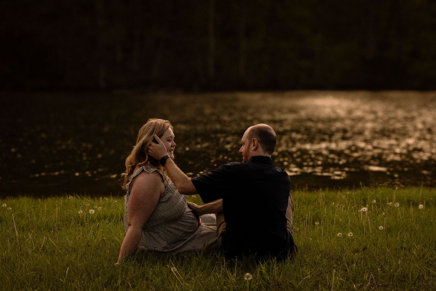 Couple relaxes on grass during sunset maternity session with dramatic lighting.