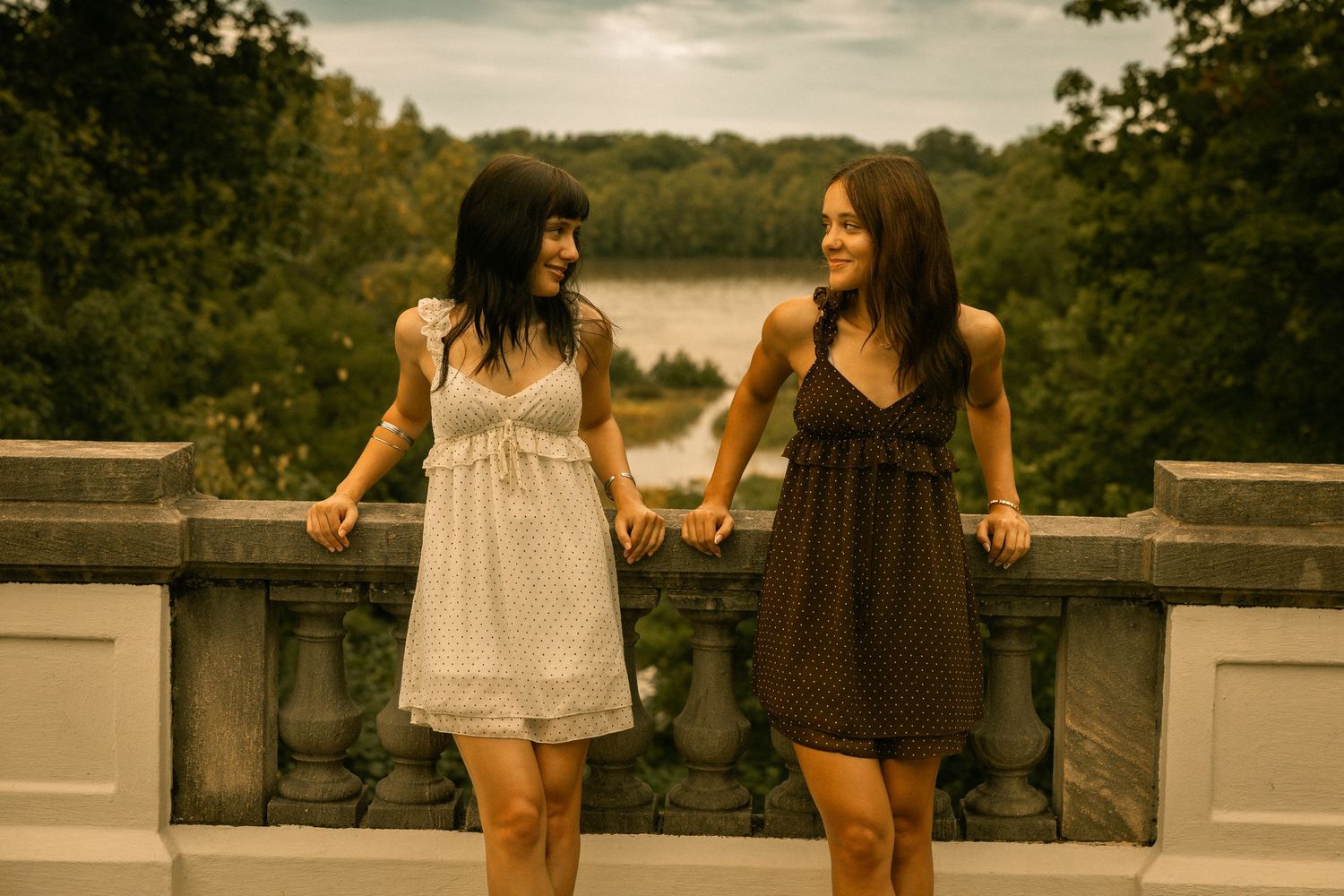 Two young women in summer dresses smile and hold hands on a stone balcony overlooking a scenic lake.