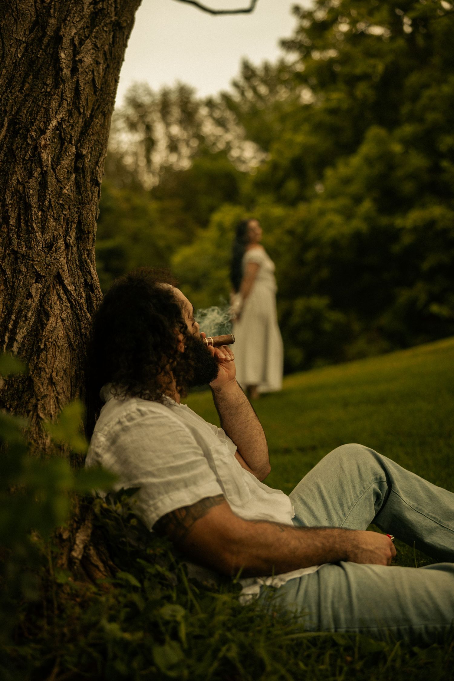 Man with curly hair resting against tree drinking wine in park, woman in white dress standing in background.