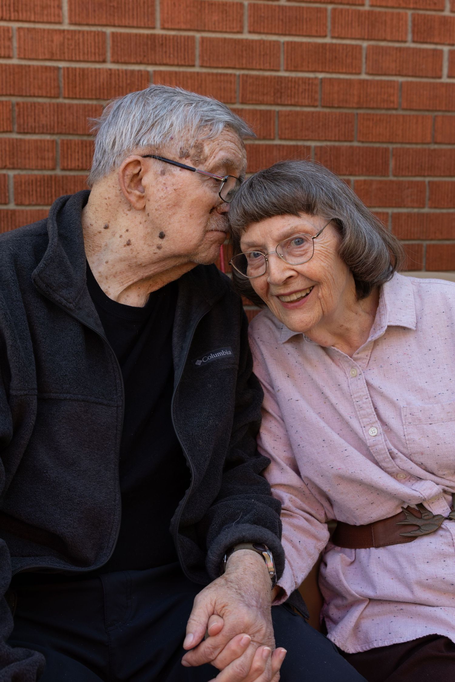 Elderly couple sharing an intimate moment while sitting together against a brick wall.