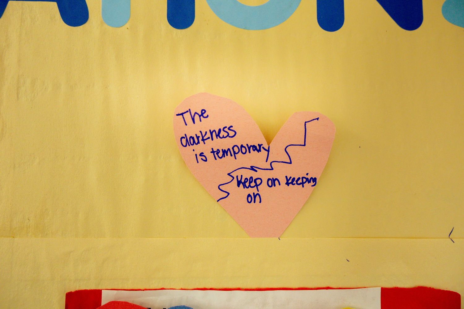 Pink heart-shaped sticky note on yellow wall with inspirational message: The darkness is temporary, keep on keeping on.