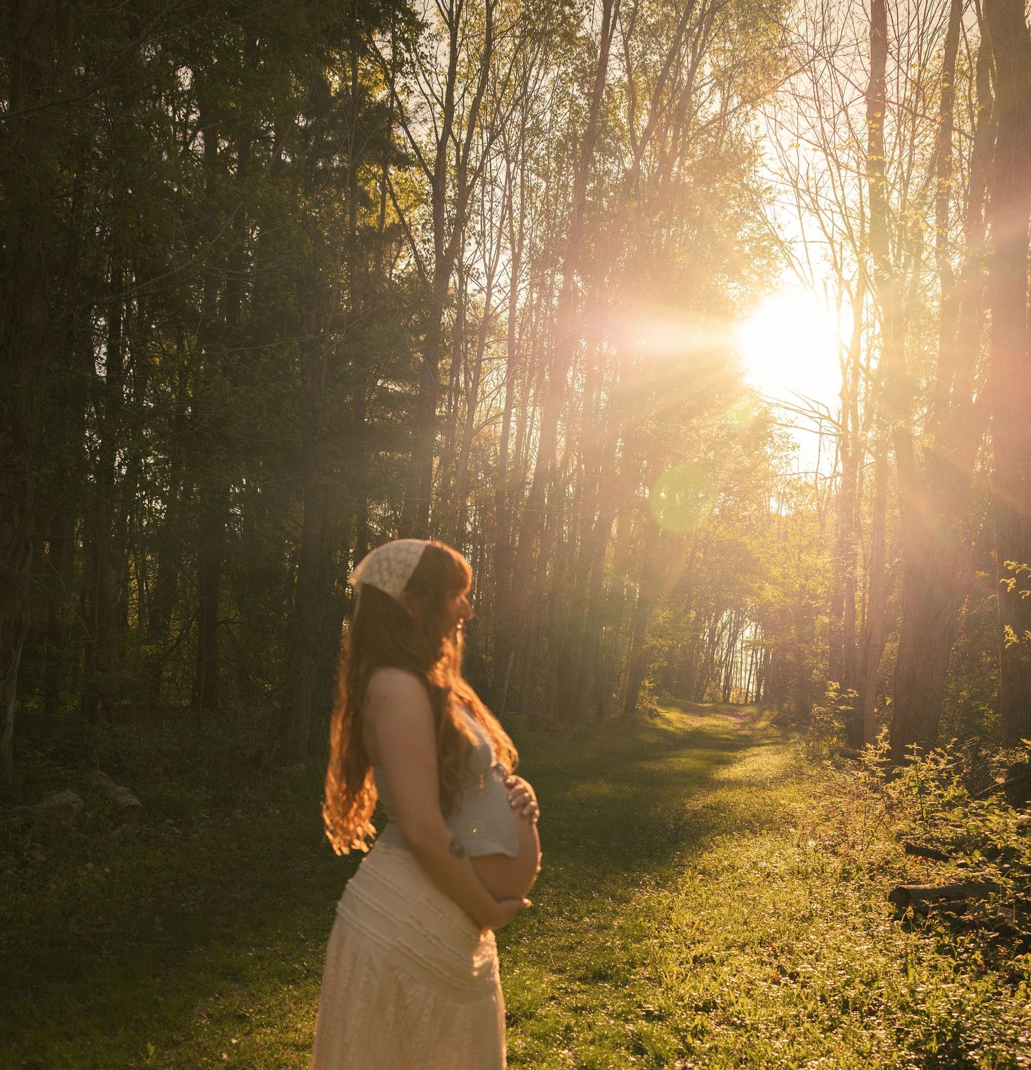 Pregnant woman in white dress holding belly in sunlit forest path during golden hour.