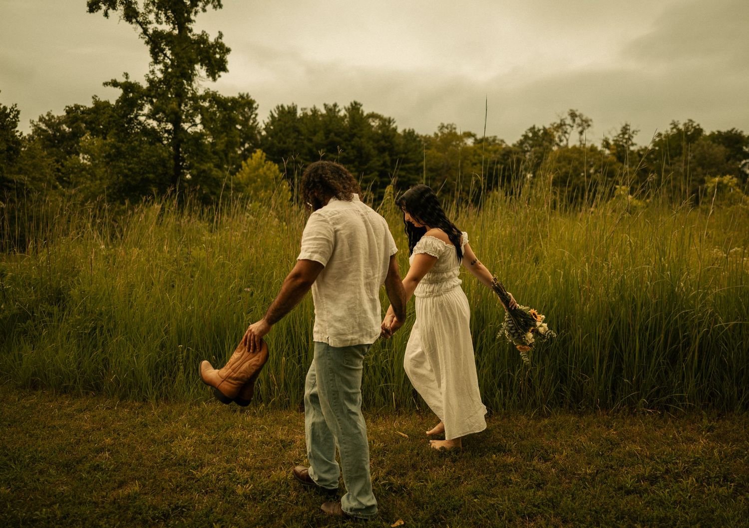 A couple walks hand-in-hand through a lush green meadow, the woman in a white dress holding wildflowers.