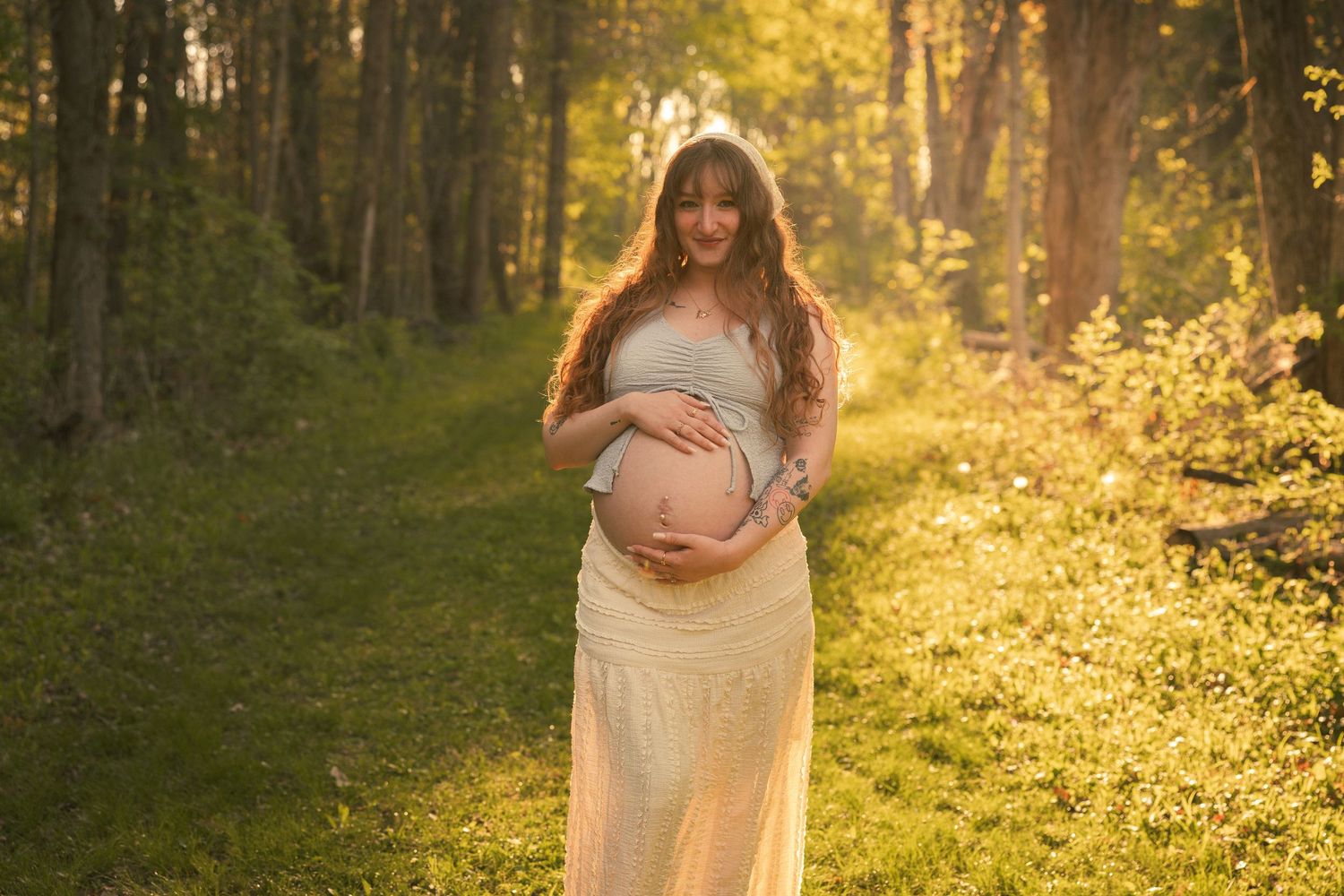 Pregnant woman in yellow dress holding baby bump in sunlit forest during golden hour maternity photoshoot.